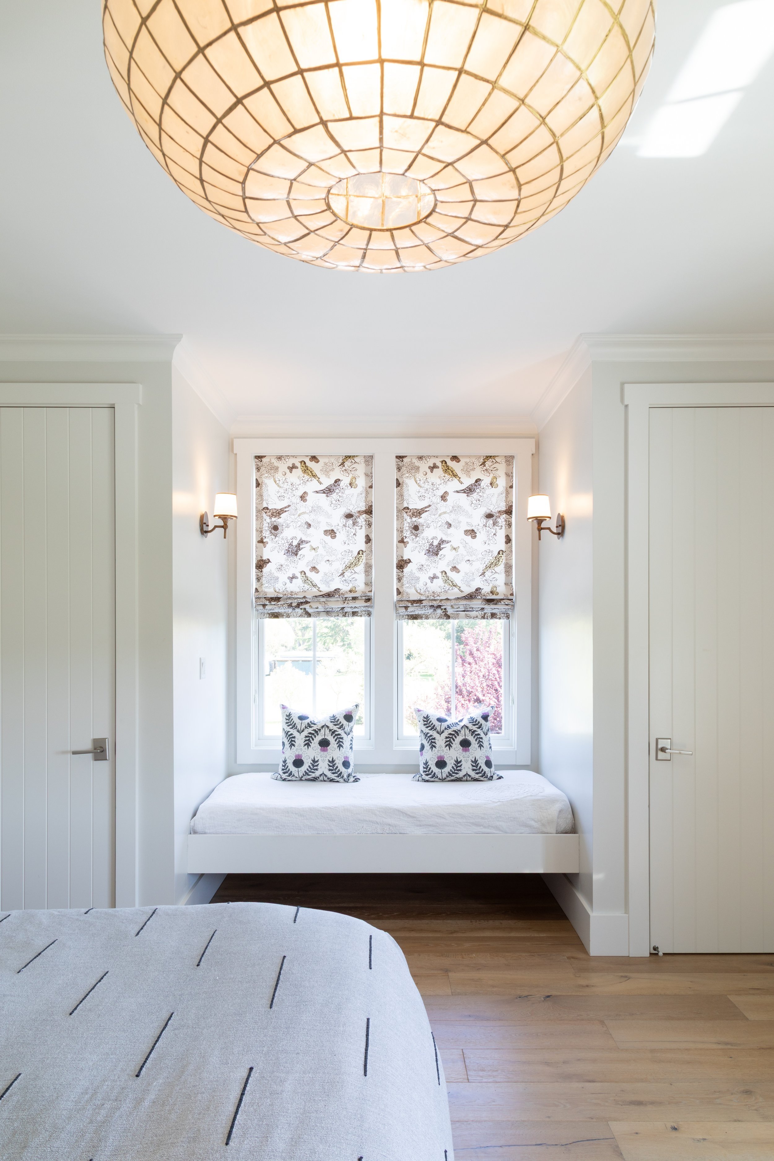 Bright bedroom with white walls, wooden floors, a large ceiling light fixture, two closed thin white doors, a window seat with two decorative pillows, and patterned window blinds.