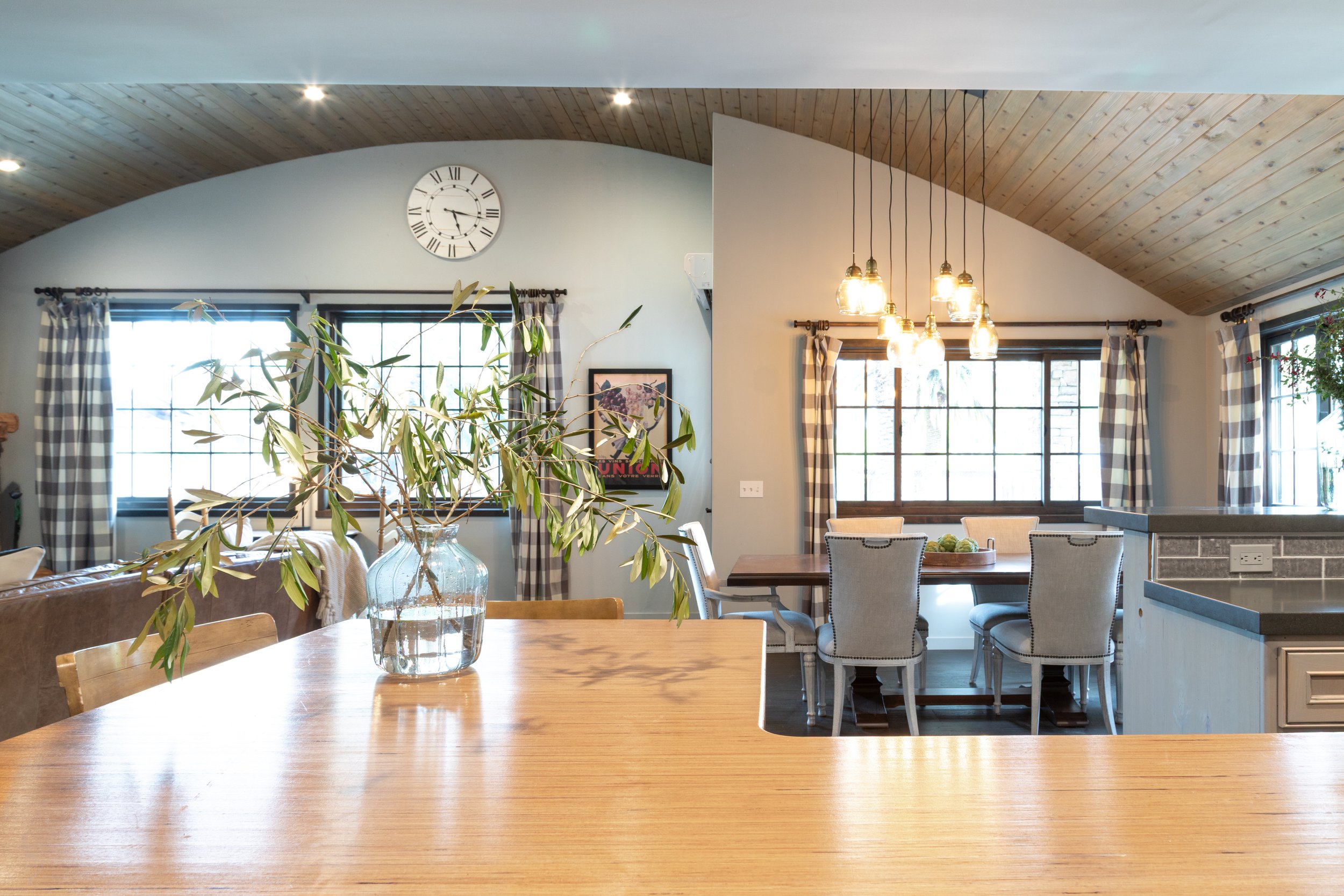 Interior of a living and dining area with wooden ceiling, checkered curtains, and a glass vase with leafy branches on a wooden table, large windows bringing in natural light, and a modern pendant light fixture above the dining table.