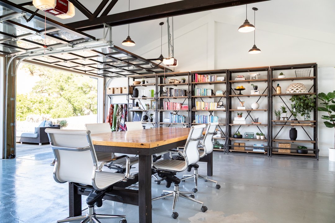 Modern garage converted into a home office with a large wooden table, white office chairs, and metal bookshelves with books and decorative items, open garage door revealing outdoor furniture and greenery.
