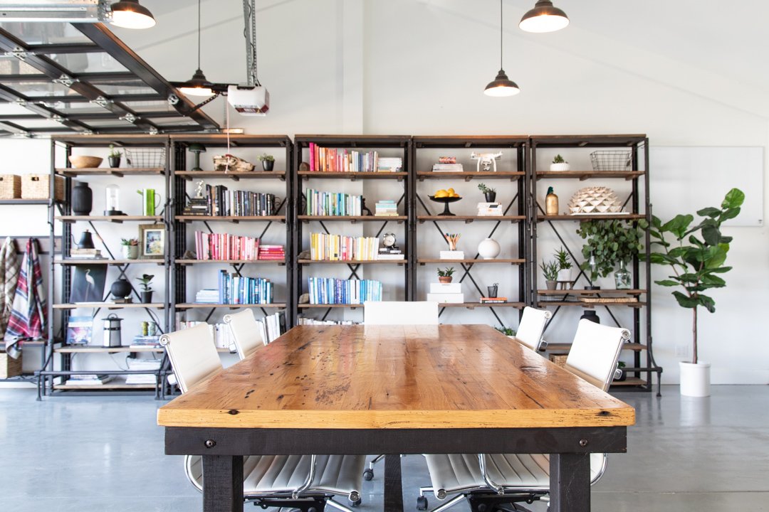 Modern conference room with a wooden table, white chairs, and metal bookshelves with books and decorative items, and a potted plant in the corner.