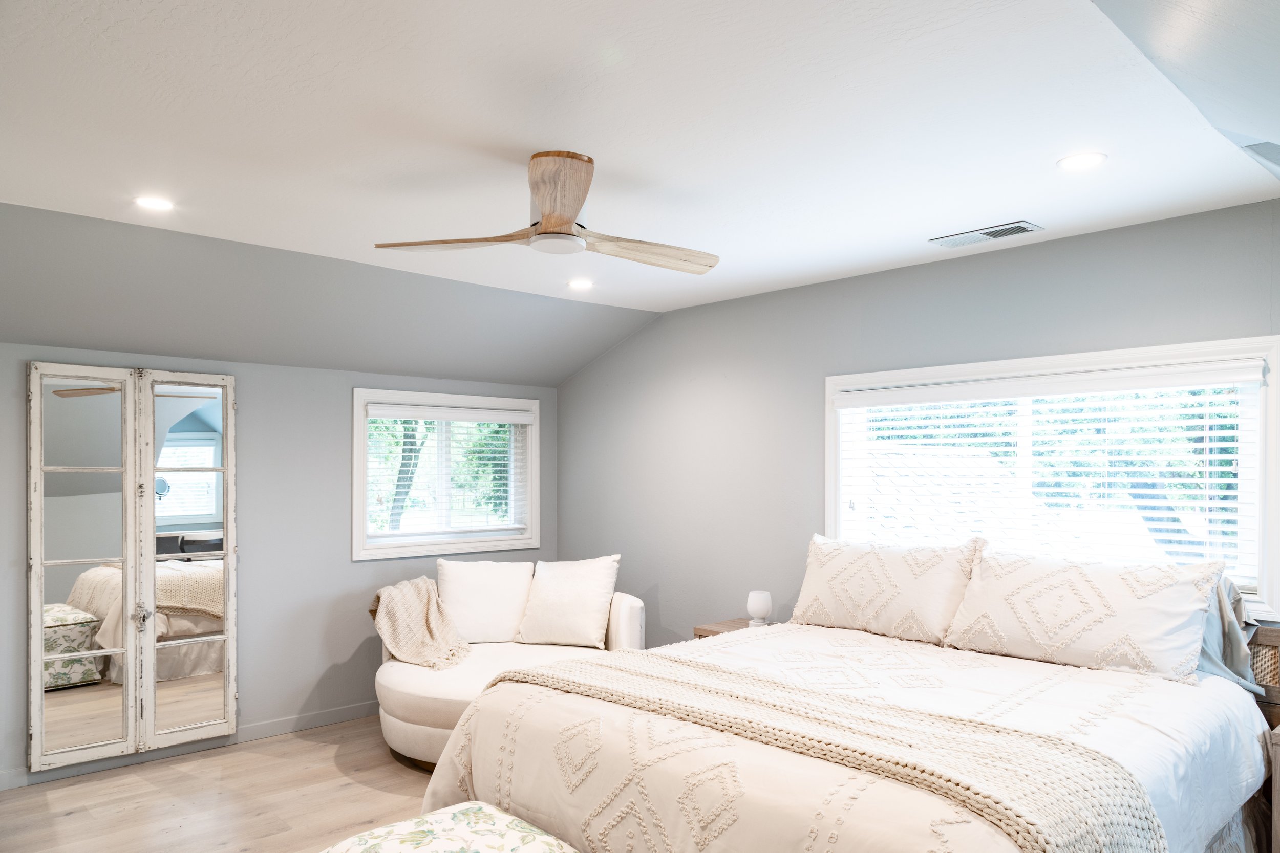 Bright bedroom with large bed, white bedding, and pillows, a small white armchair, two windows with blinds, a white distressed full-length mirror, light wood flooring, and a ceiling fan with wooden blades.