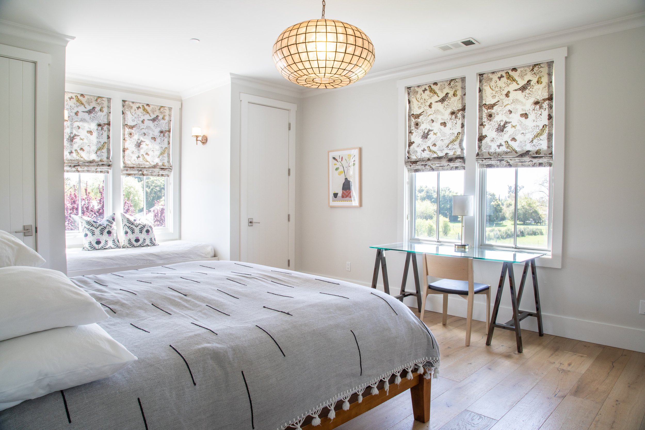 A bright, modern bedroom with a large bed, a window seat with patterned cushions, a desk with a chair, and large windows with bird-patterned roman shades.