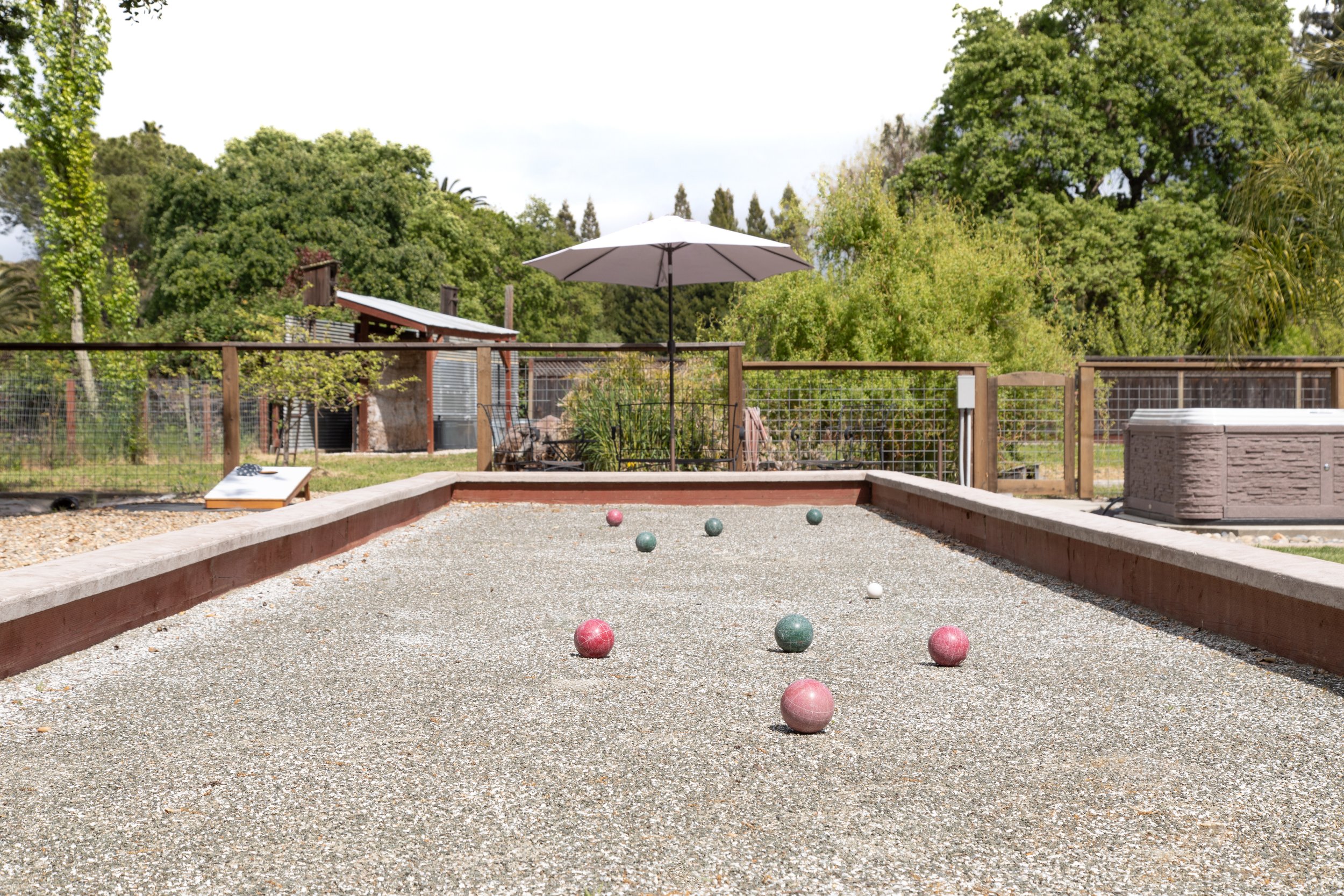 Outdoor bocce ball court with colored balls scattered on the gravel surface, surrounded by a wooden border, with trees and a fence in the background, and an umbrella providing shade.