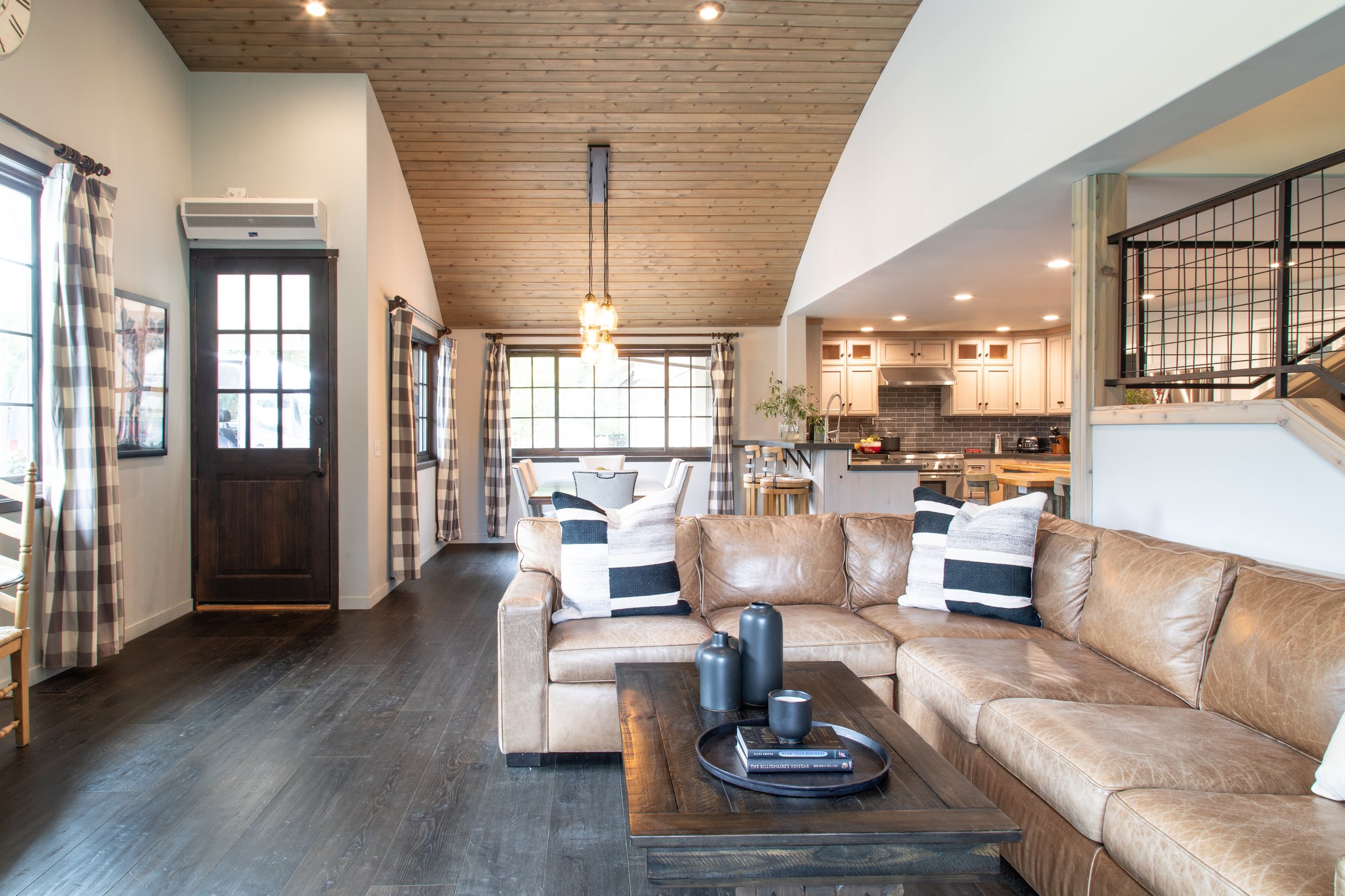 Living room with tan leather sectional sofa, dark wood coffee table, black and white striped pillows, wood and black metal railing, kitchen with light cabinets and black backsplash, large front window, dining table with chairs, wood vaulted ceiling,
