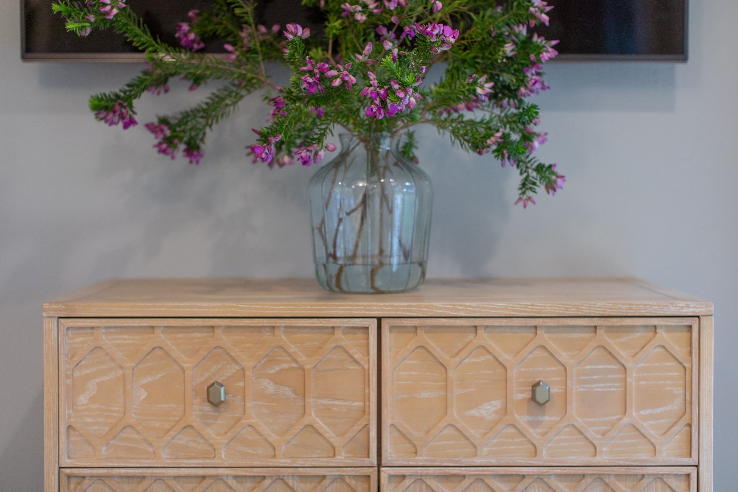 A wooden dresser with a geometric pattern on the drawer fronts, topped with a clear glass vase holding purple and pink flowers.