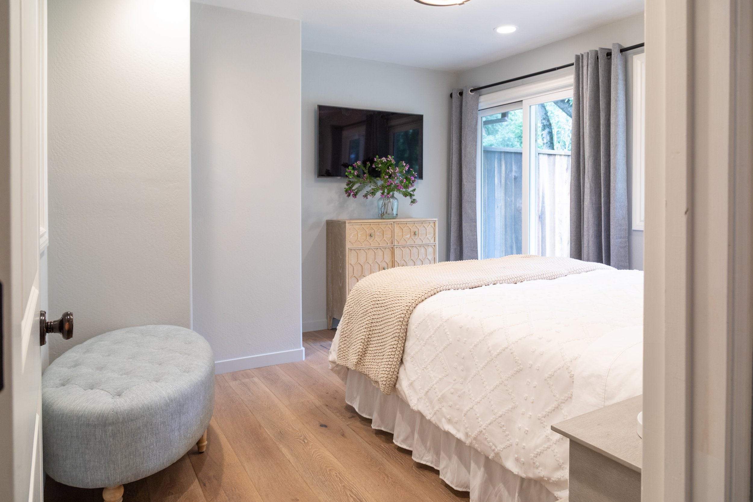 Modern bedroom with white bedding, wooden flooring, sliding glass door with gray curtains, wall-mounted TV, wooden dresser with a vase of purple flowers, and a small upholstered bench.
