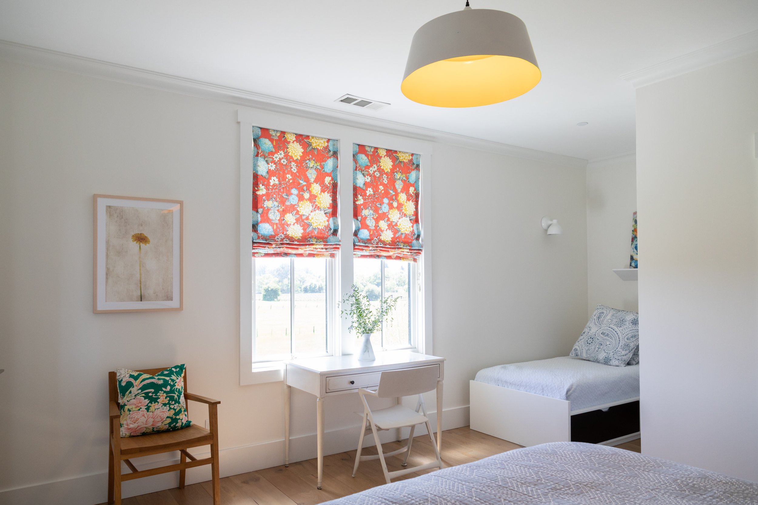 Bright bedroom with twin bed, white walls, floral window shades, small desk and chair, framed art, and a wooden chair with floral cushion.
