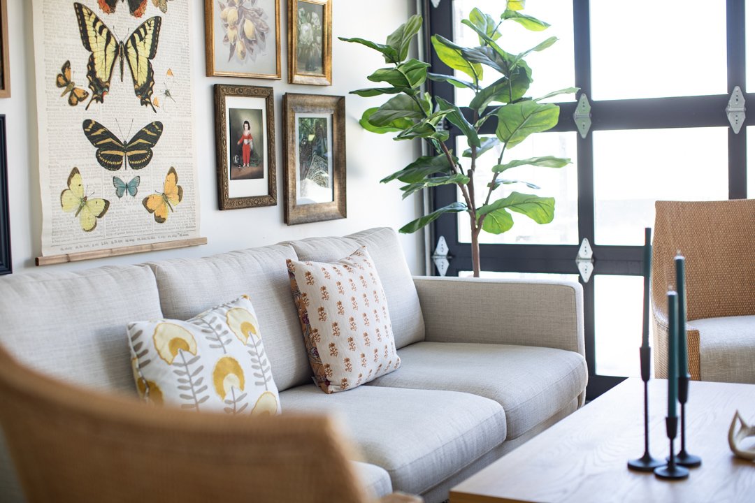 Living room with a beige sofa, patterned pillows, large green potted plant, wall art including butterflies and framed pictures, black-framed window, and a wooden table with candles.