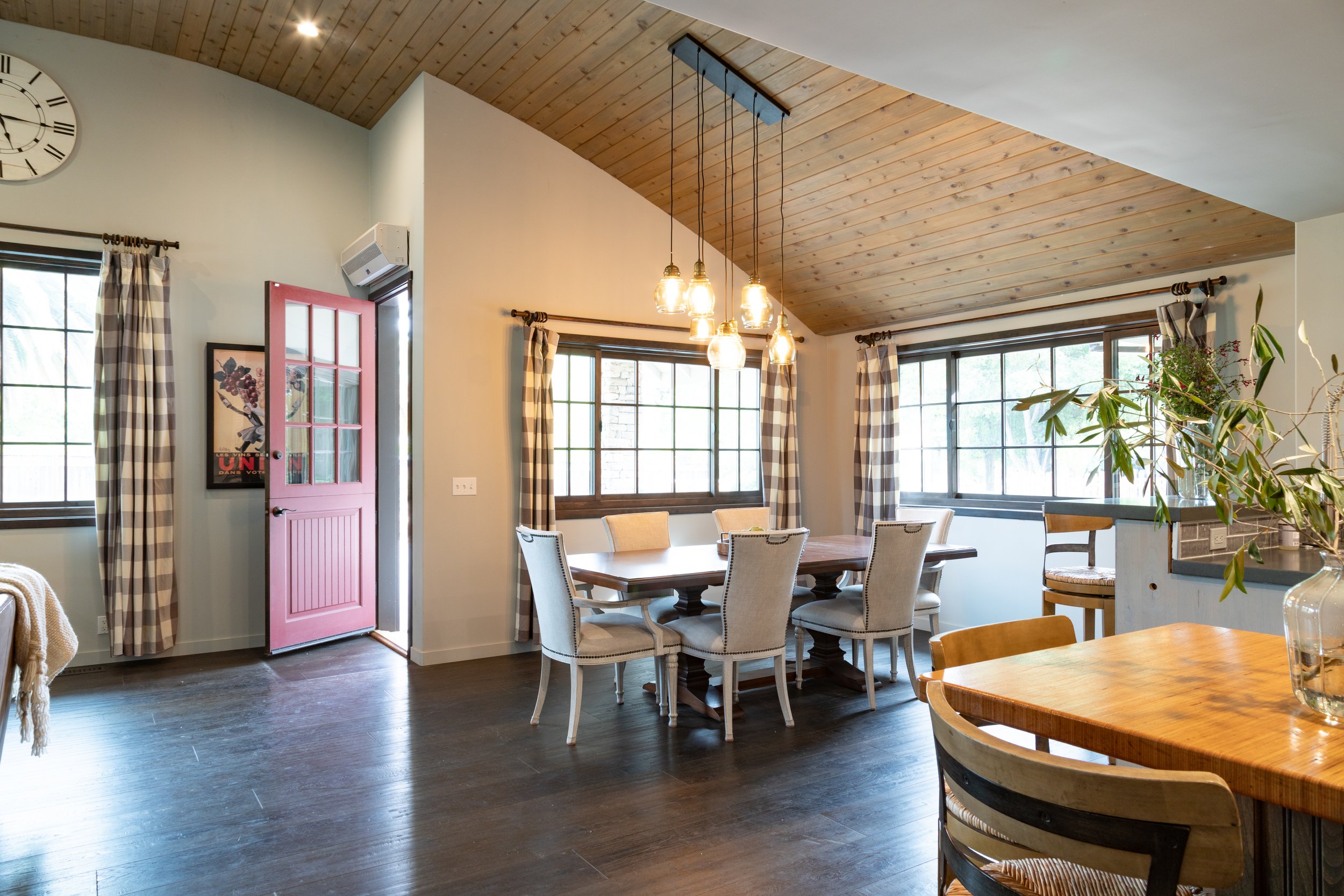 Bright dining room with wooden paneled ceiling, large windows with plaid curtains, a central table surrounded by white upholstered chairs, and a smaller table with a vase of greenery.