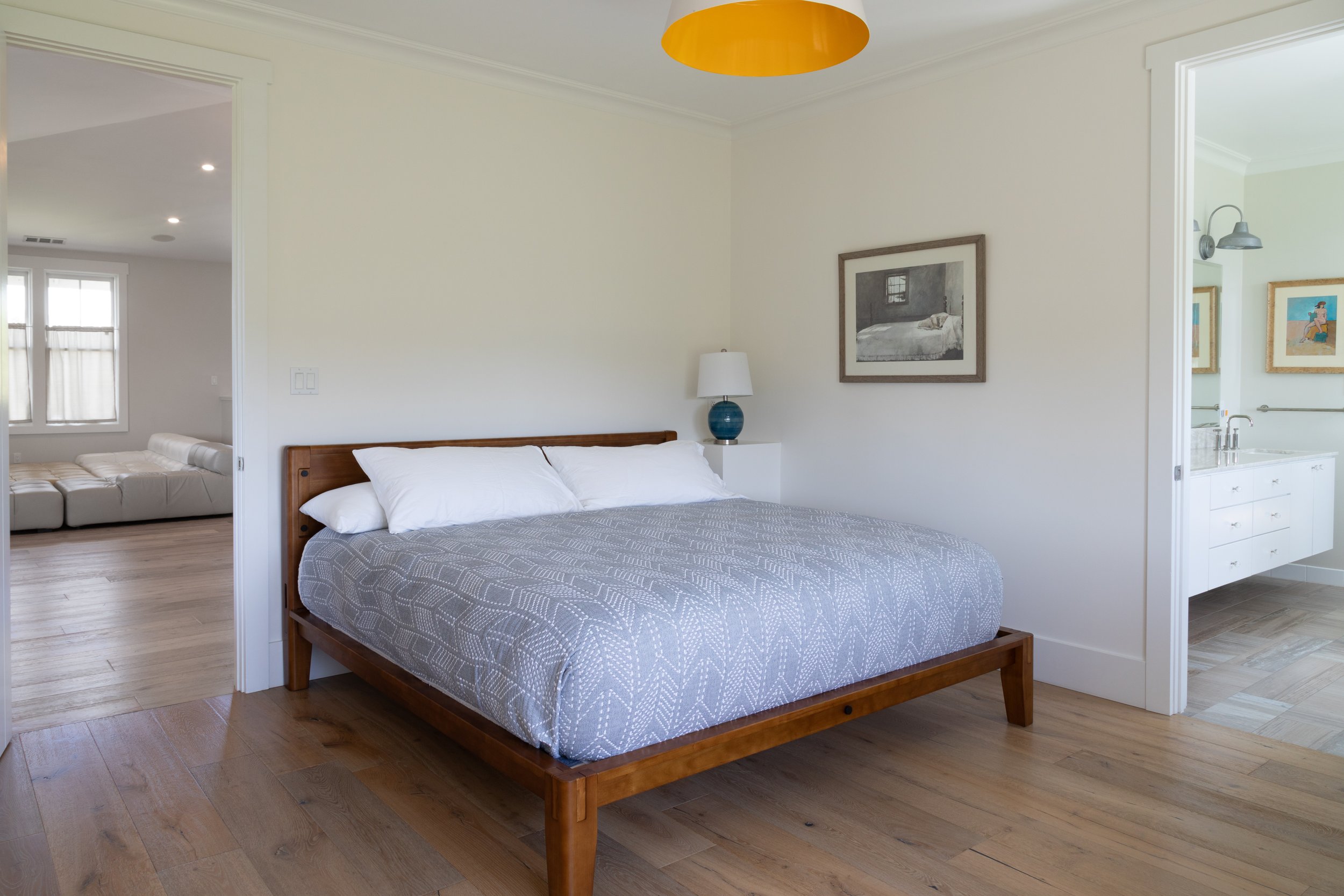 A bedroom with a wooden bed frame, white pillows, grey patterned bedding, a white nightstand with a blue lamp, a framed black and white photograph, and a poster on the wall. An open doorway leads to a sitting area with a white leather couch, and anot
