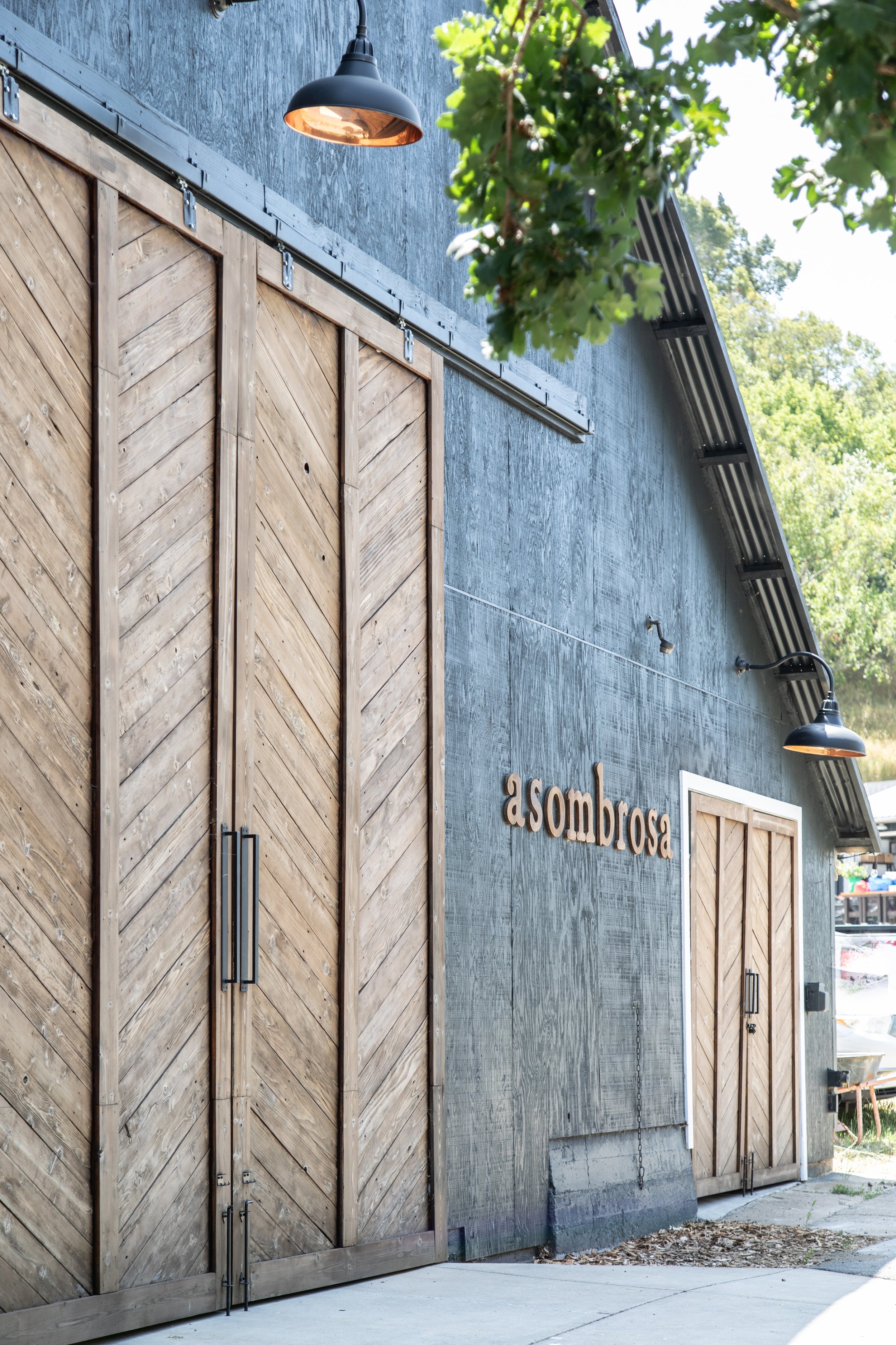Exterior of a building with wooden barn-style sliding doors, black outdoor lights, and a sign that says "a sombrosa" mounted on a gray textured wall. Green leaves partially visible at the top.