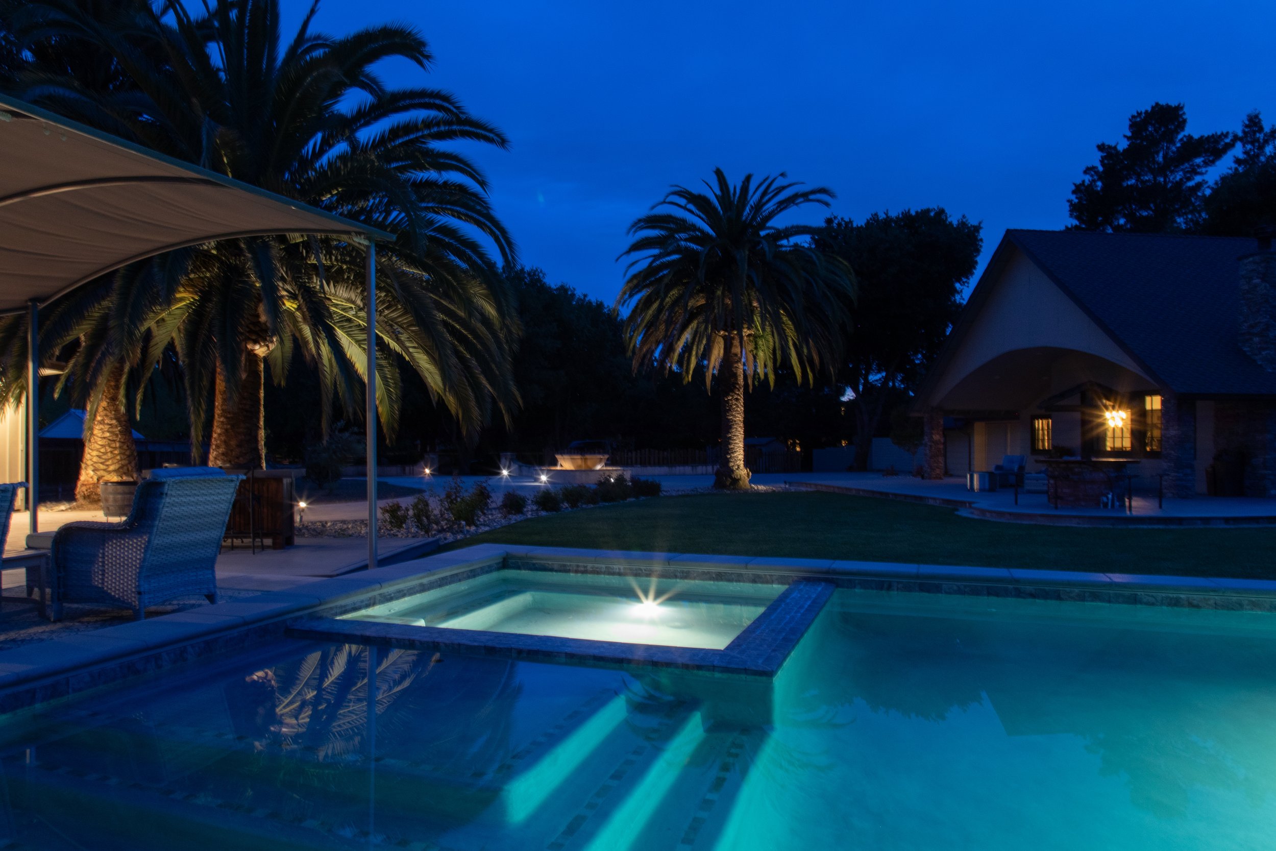 A backyard at night with illuminated swimming pool, palm trees, patio furniture, and a house with lights on.