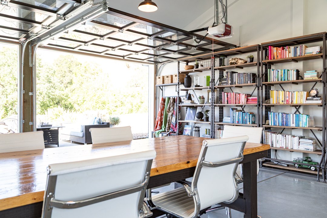 Modern garage converted into a home office with a large wooden table and white chairs, metal shelving filled with books and decor, and an open garage door revealing a green outdoor space.