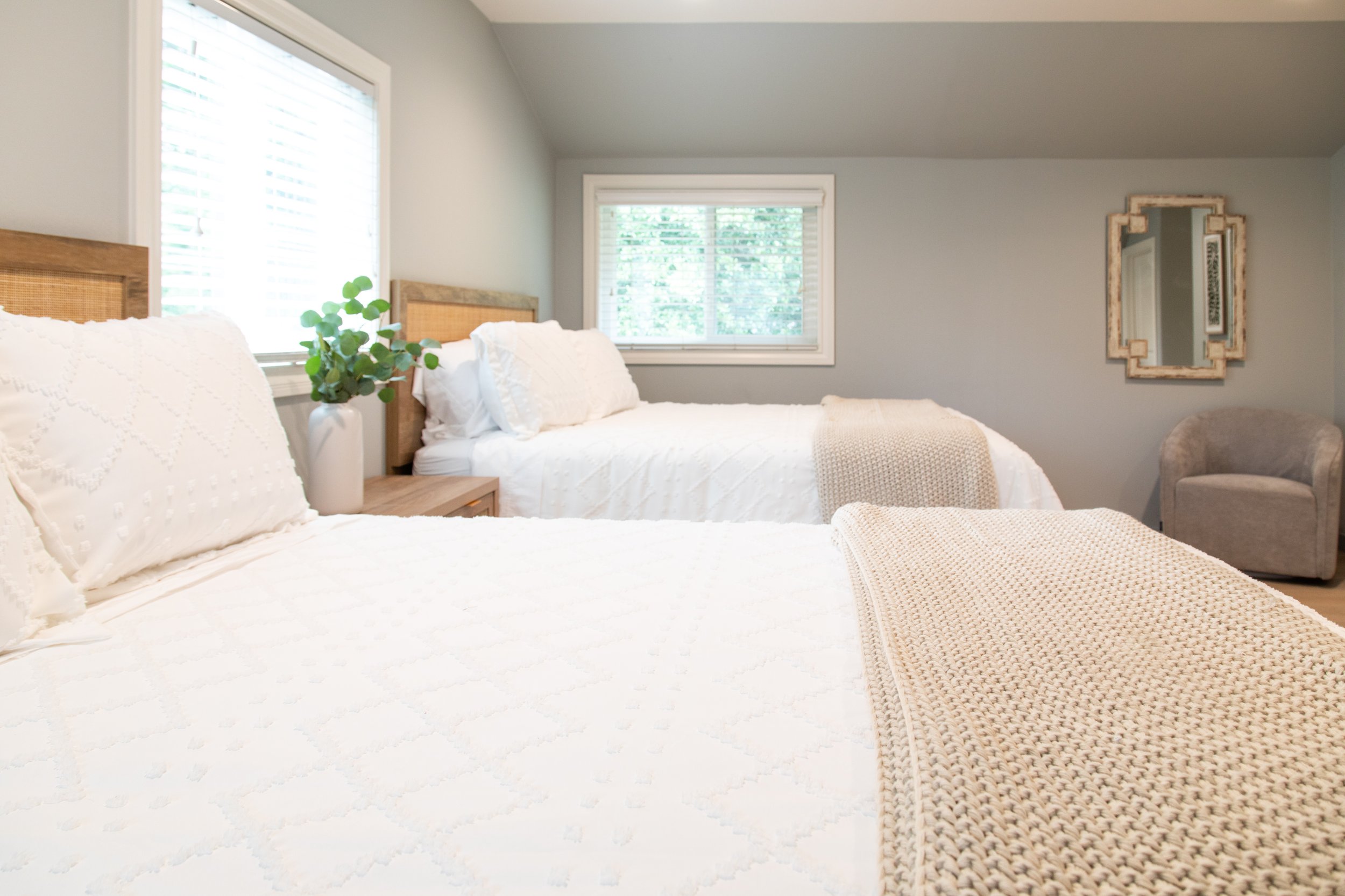 A cozy bedroom with two beds, white bedding, decorative knitted blankets, a side table with a plant, and a mirror on the wall.