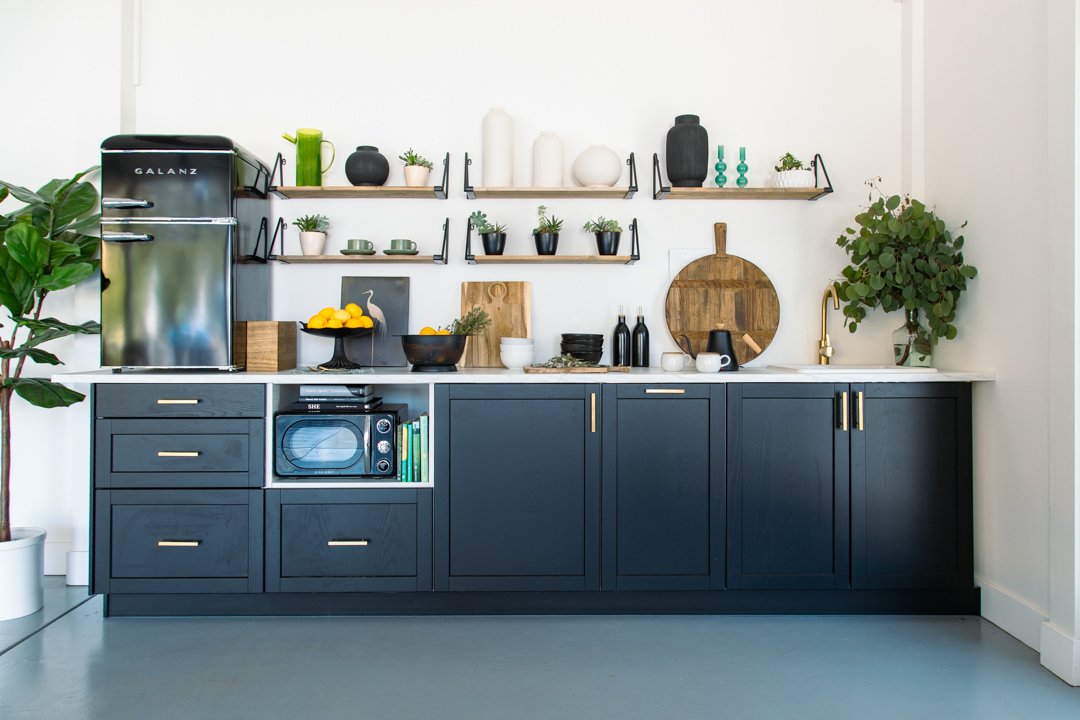 Modern kitchen with dark blue cabinets, open shelves with various pots and vases, microwave, small refrigerator, and potted plants.