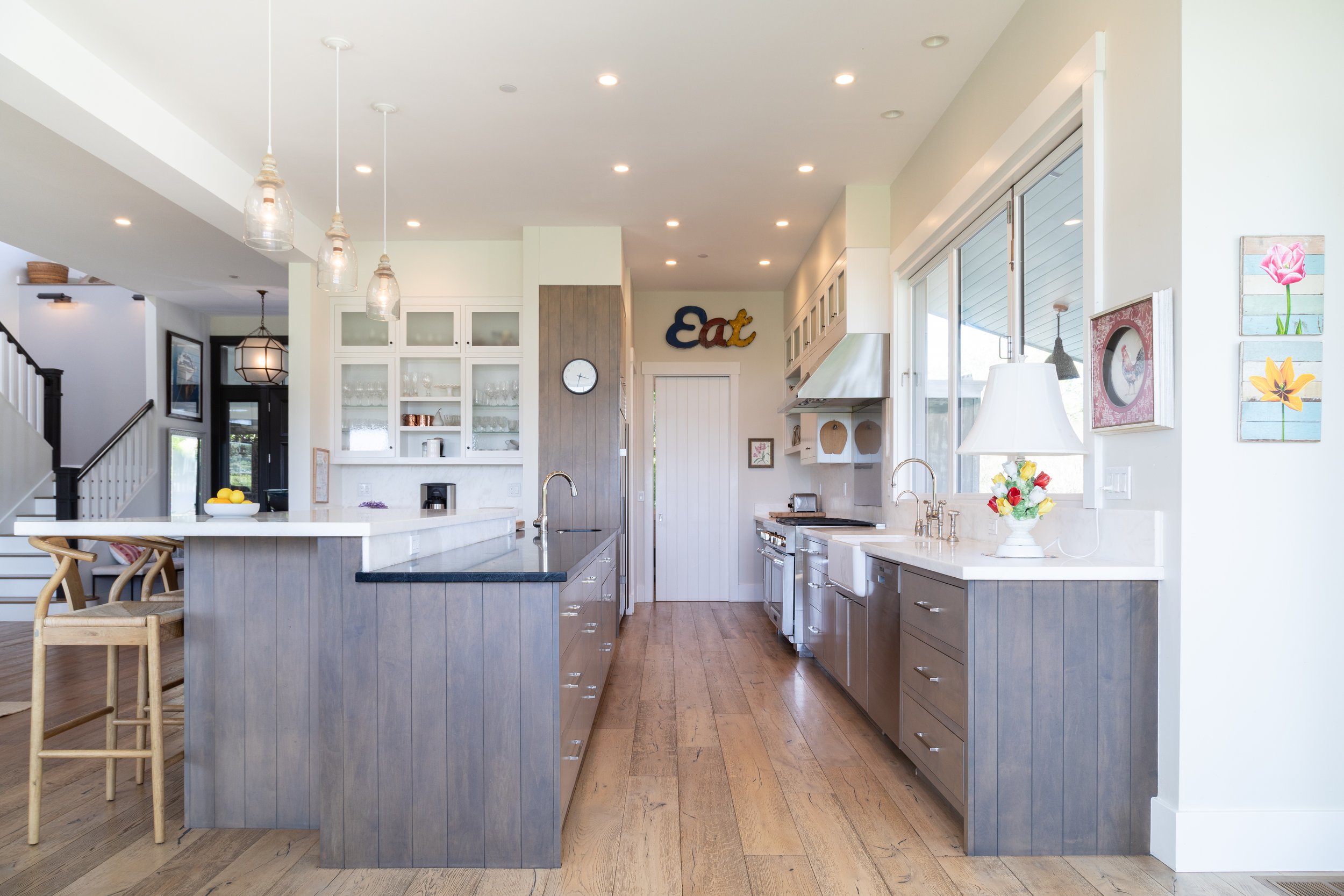 Modern kitchen with white and gray cabinets, wooden flooring, and large windows. Decor includes colorful artwork, a vase with flowers, and a sign reading 'Eat' above a door.