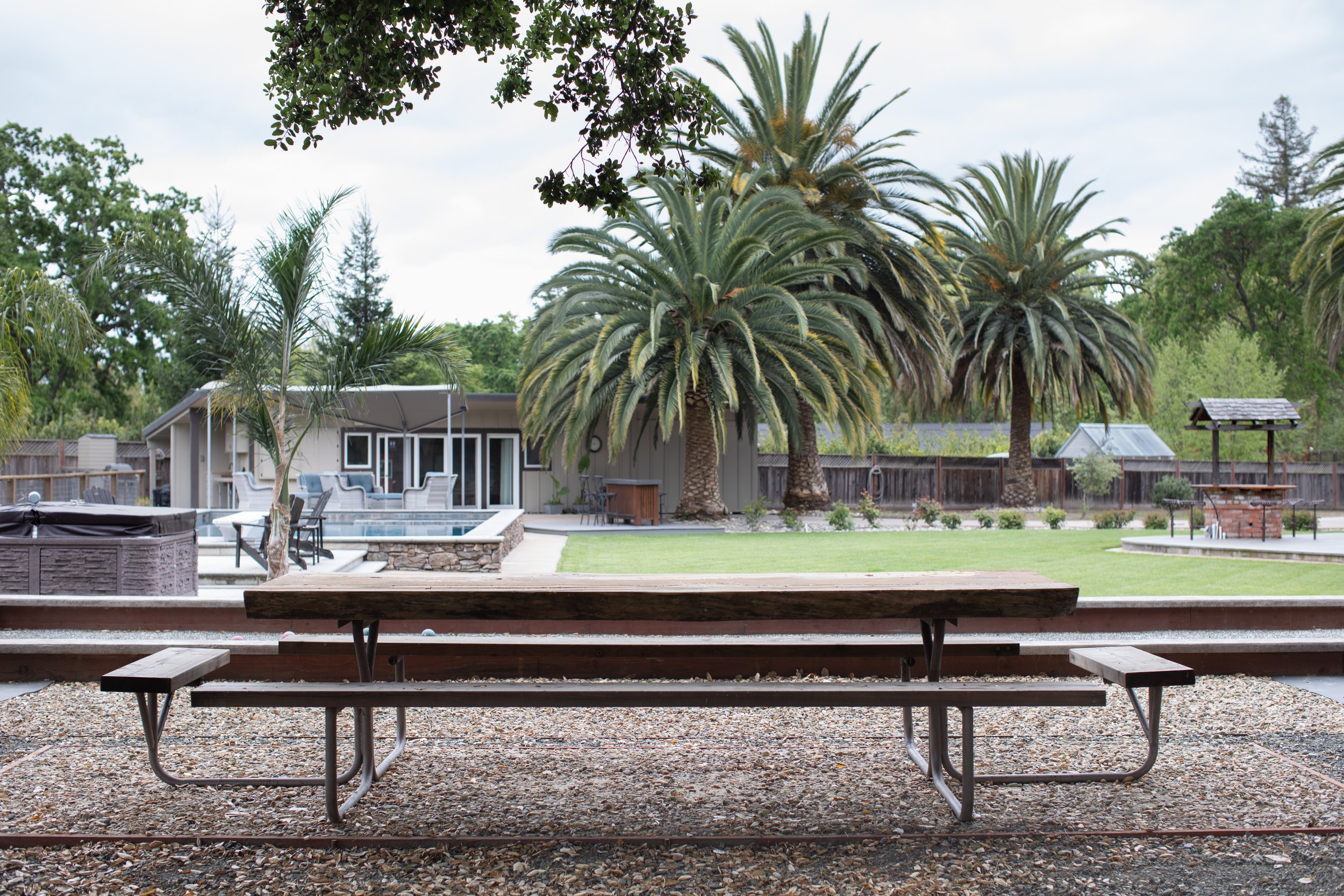 A backyard view from a covered patio with a wooden picnic table and benches, overlooking a landscaped yard with palm trees, a small pool, outdoor seating, and a brick barbecue grill.