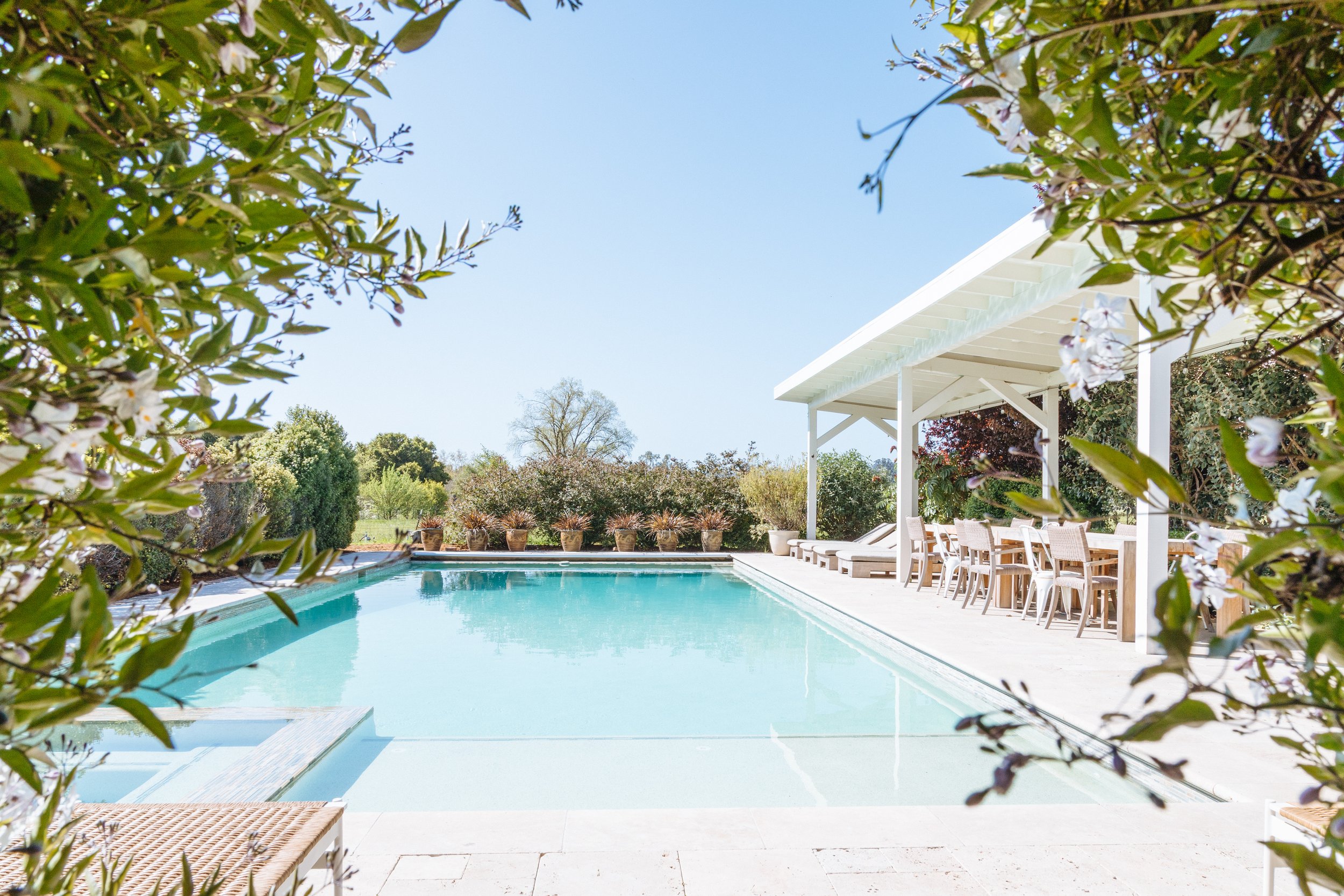View of a backyard pool with surrounding lounge chairs, potted plants, and a white shaded patio area in a sunny, open yard.