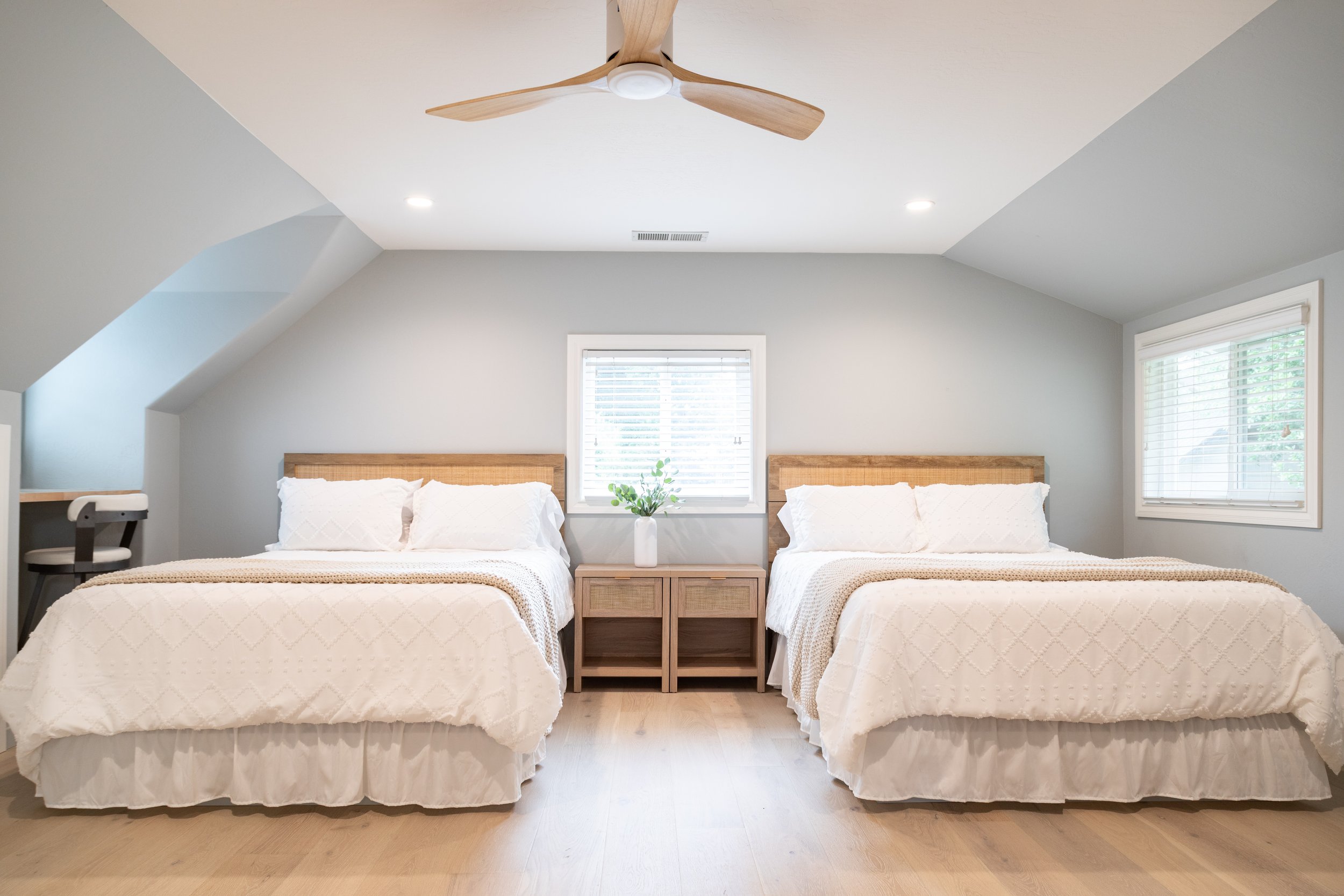 A bedroom with two beds, each with white bedding, wooden headboards, and a nightstand with a vase and plant between them. The room has white walls, windows with blinds, a ceiling fan, and recessed lighting.