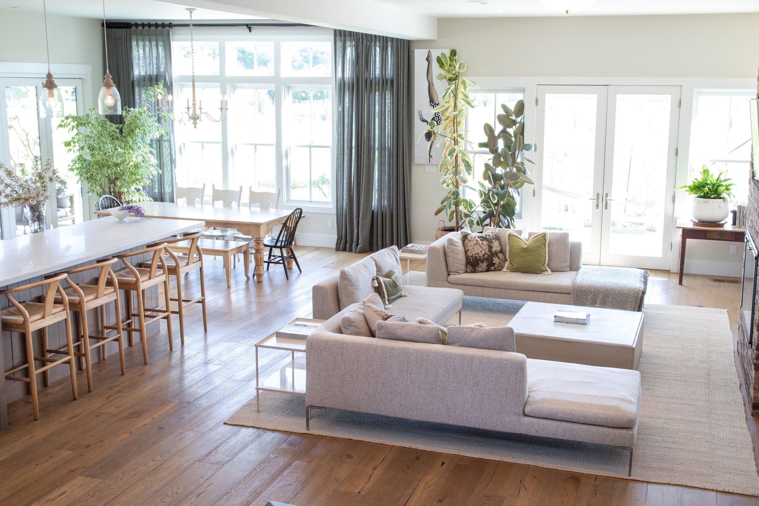 Bright living room with beige sofas, large windows with dark curtains, potted plants, wooden flooring, and a dining area with a white table and chairs.