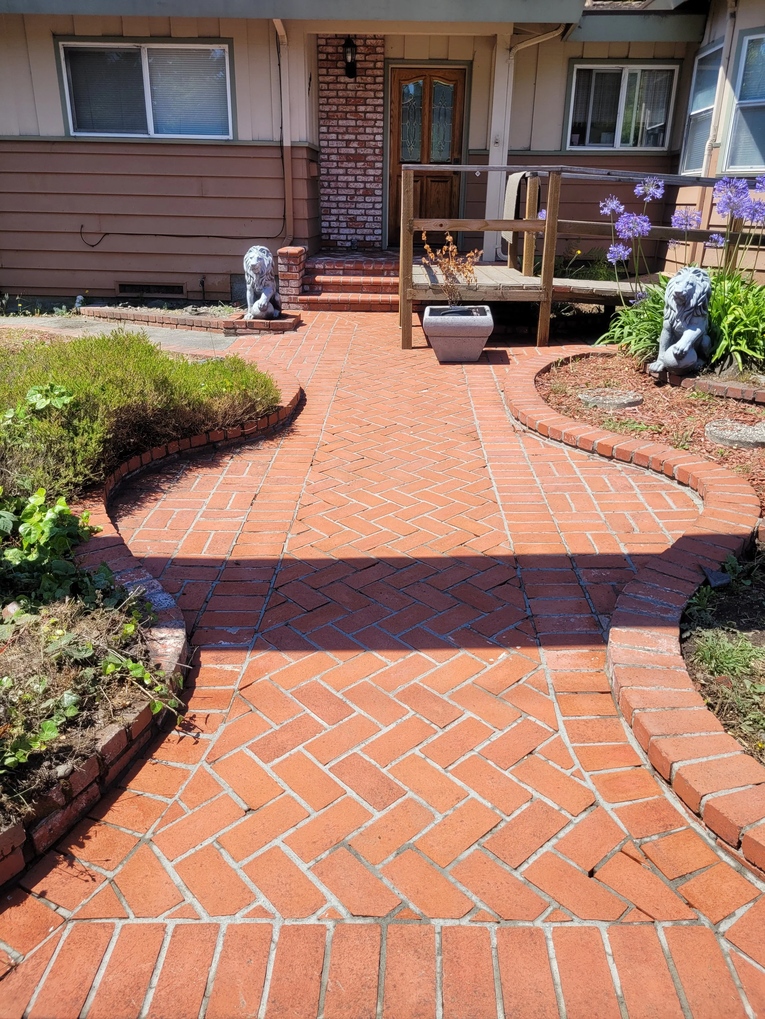 Front entrance of house with brick walkway, two stone lion statues, and garden areas with plants and purple flowers.
