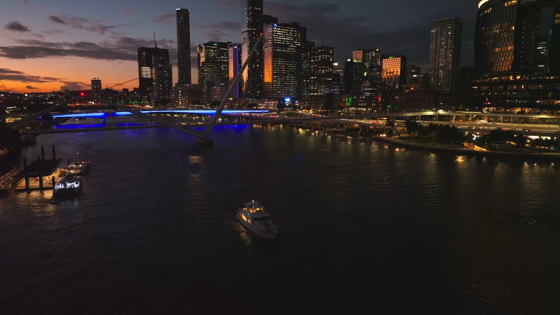 Nighttime view of a city skyline over a river with illuminated buildings, a bridge, and boats on the water.