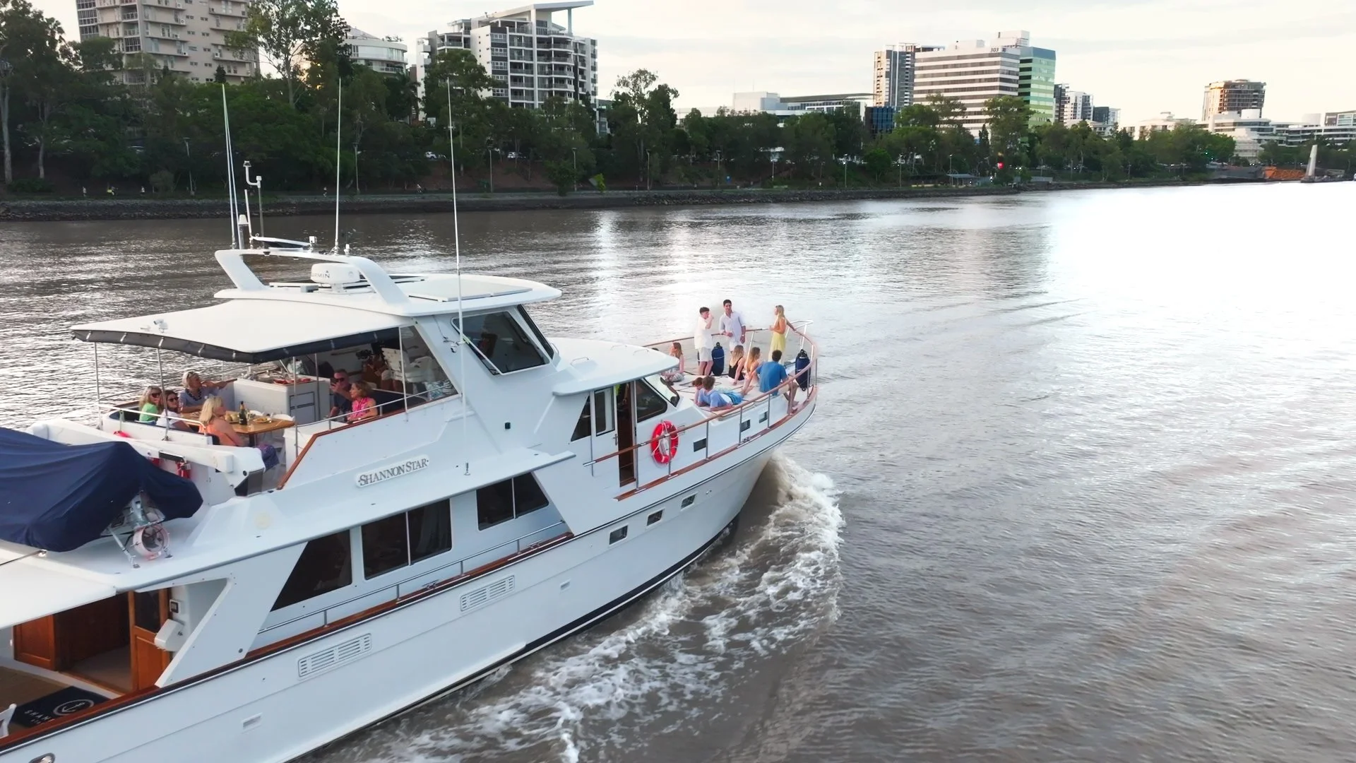 A white yacht named 'Shannon Star' with people onboard, sailing on the Brisbane River with the city skyline of modern buildings in the background.
