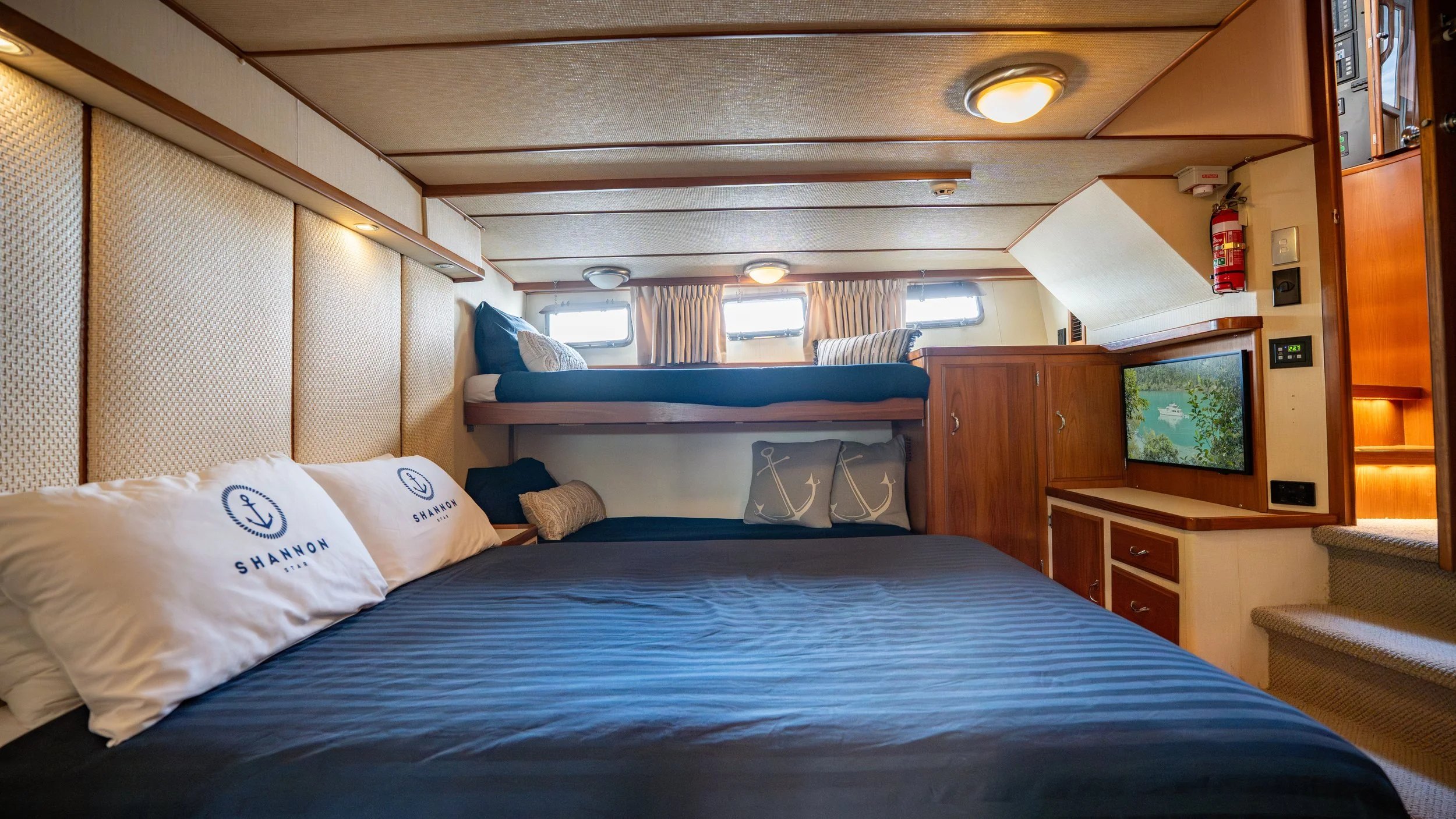 Interior of a boat cabin with a large bed, pillows with anchor and shell designs, a shorter bed, three small windows with curtains, a backdrop of wood paneling, and a TV screen showing a water scene.