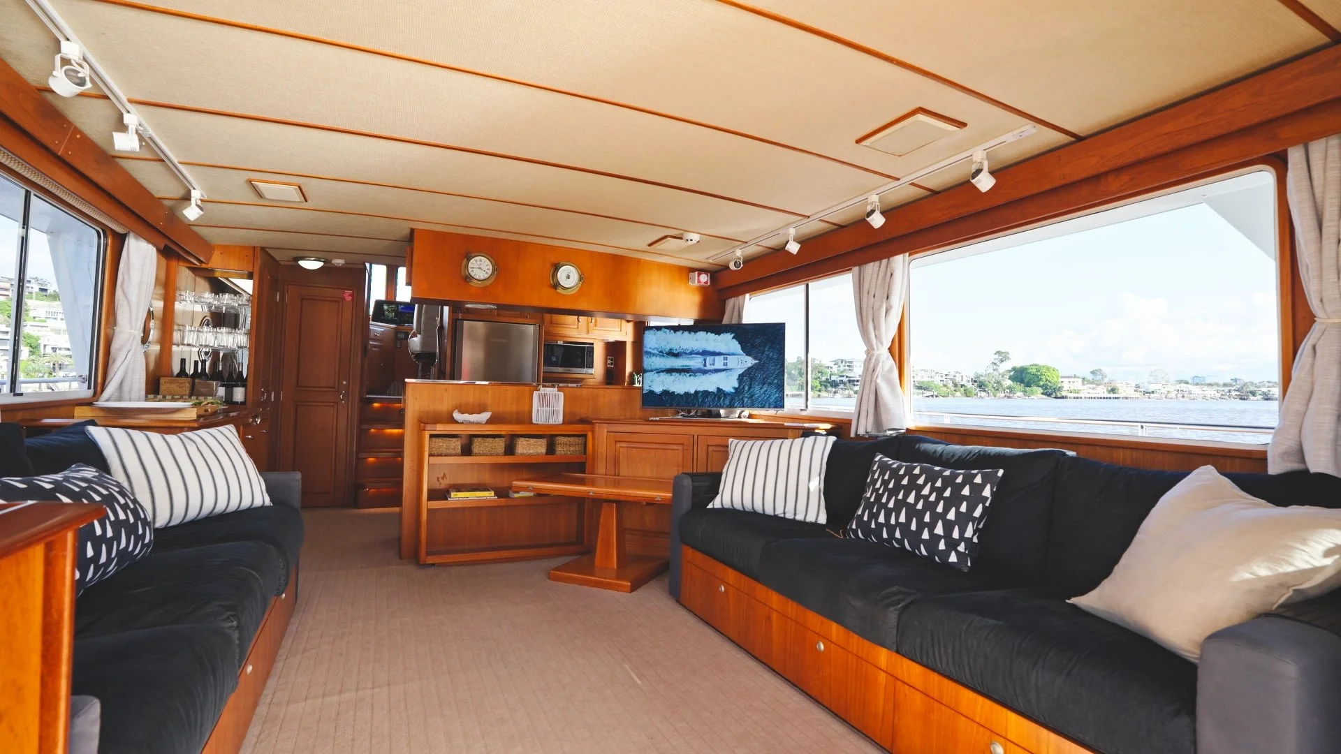 Interior of a boat or yacht living area with wooden paneling, black sofas with striped and patterned pillows, a wall-mounted TV, large windows showing water and shoreline, and various small decorative items.
