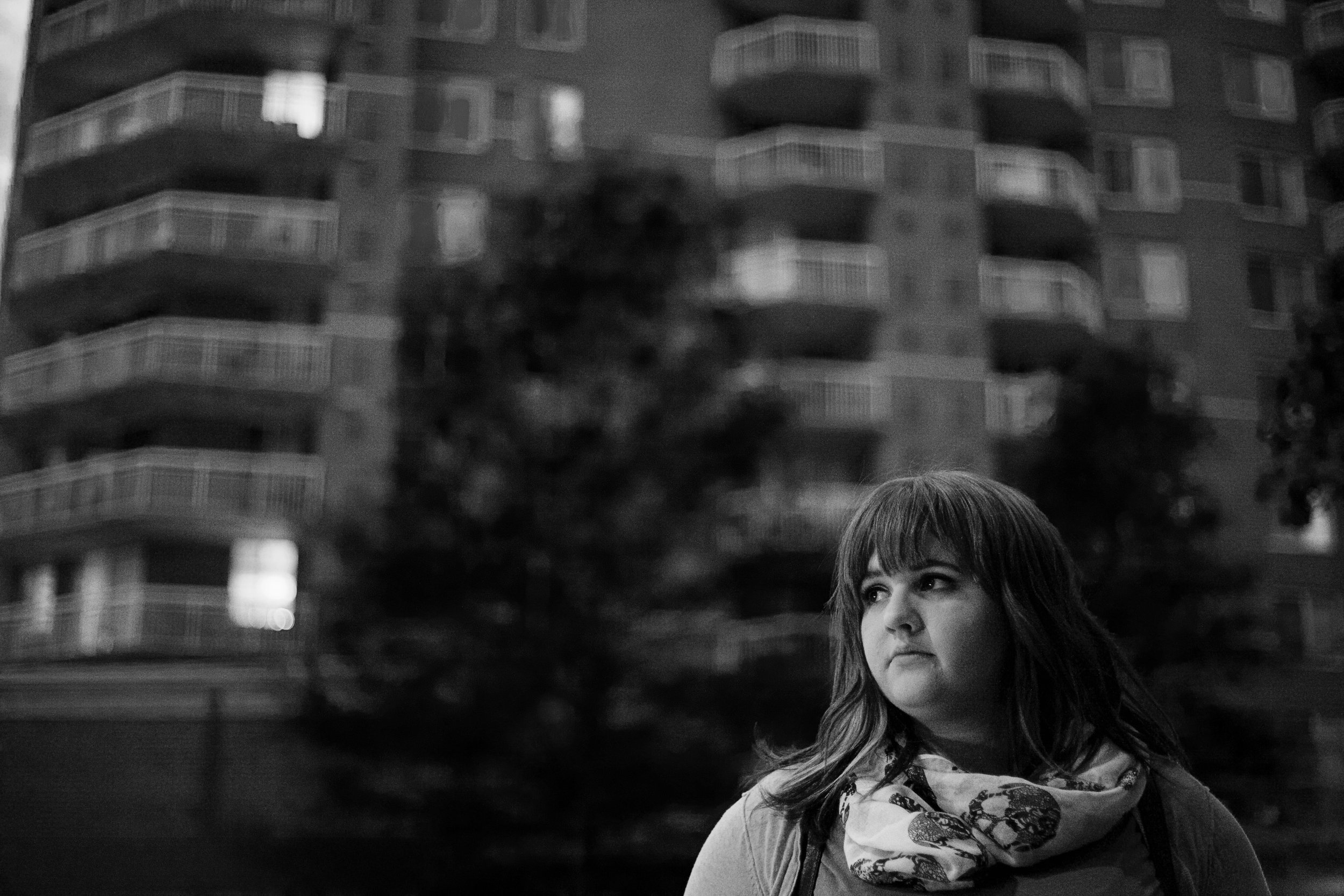 A woman with shoulder-length hair wearing a scarf around her neck standing outdoors in front of a blurred apartment building.