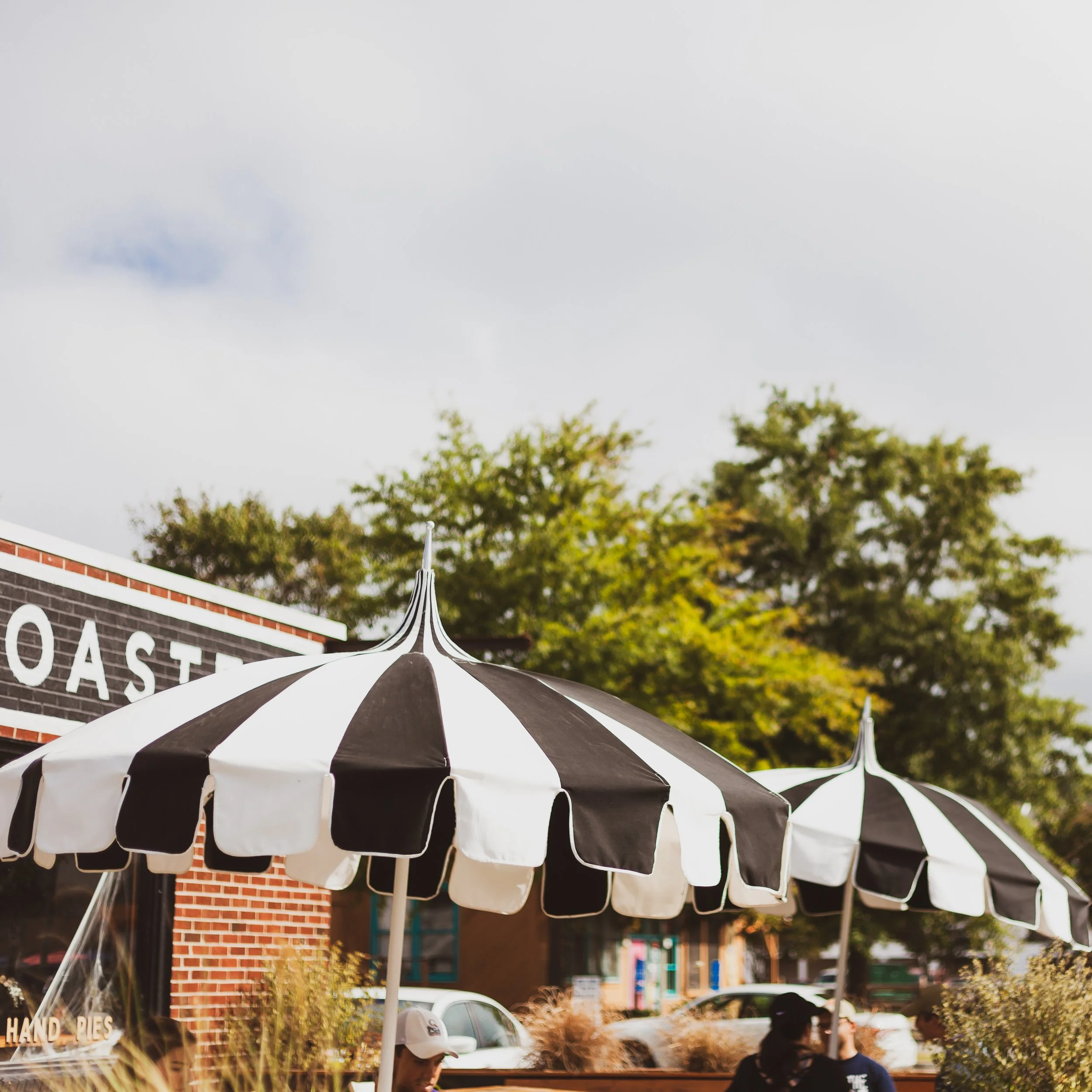 Black and white striped outdoor umbrellas in front of a brick building with people walking by and trees in the background.
