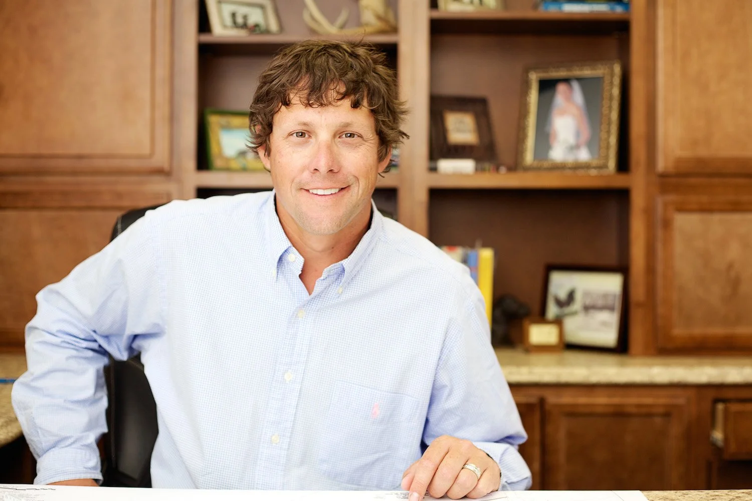 A man with brown, wavy hair and light skin, smiling while sitting at a desk in a construction office with wooden shelves in the background containing framed photographs and books.
