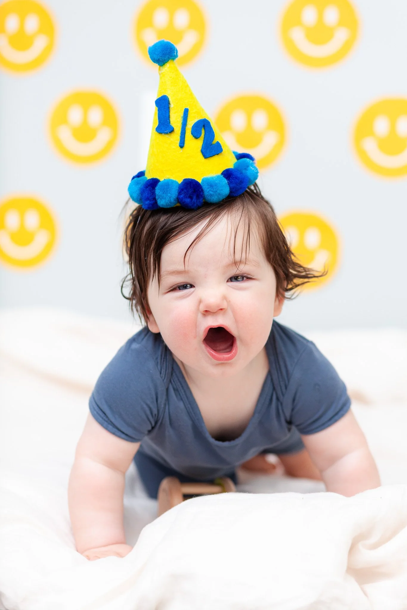 A bright, colorful, and airy shot of an infant in a blue shirt and yellow birthday hat with blue pom-poms and the number 1/2 happily shouts at the camera on a white blanket in front of a yellow smiley face background.