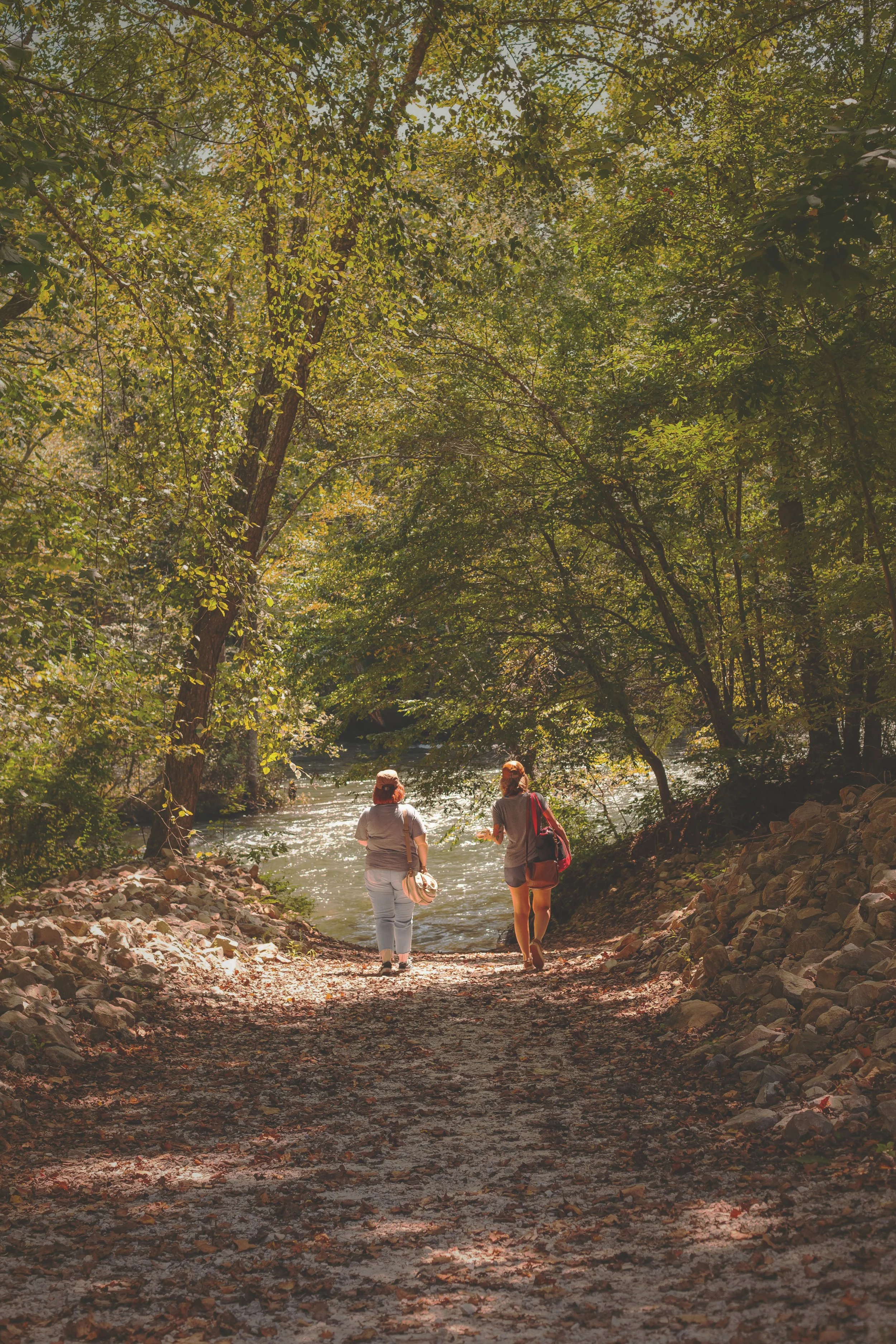 Two women walking along a dirt trail near a river surrounded by dense green trees.