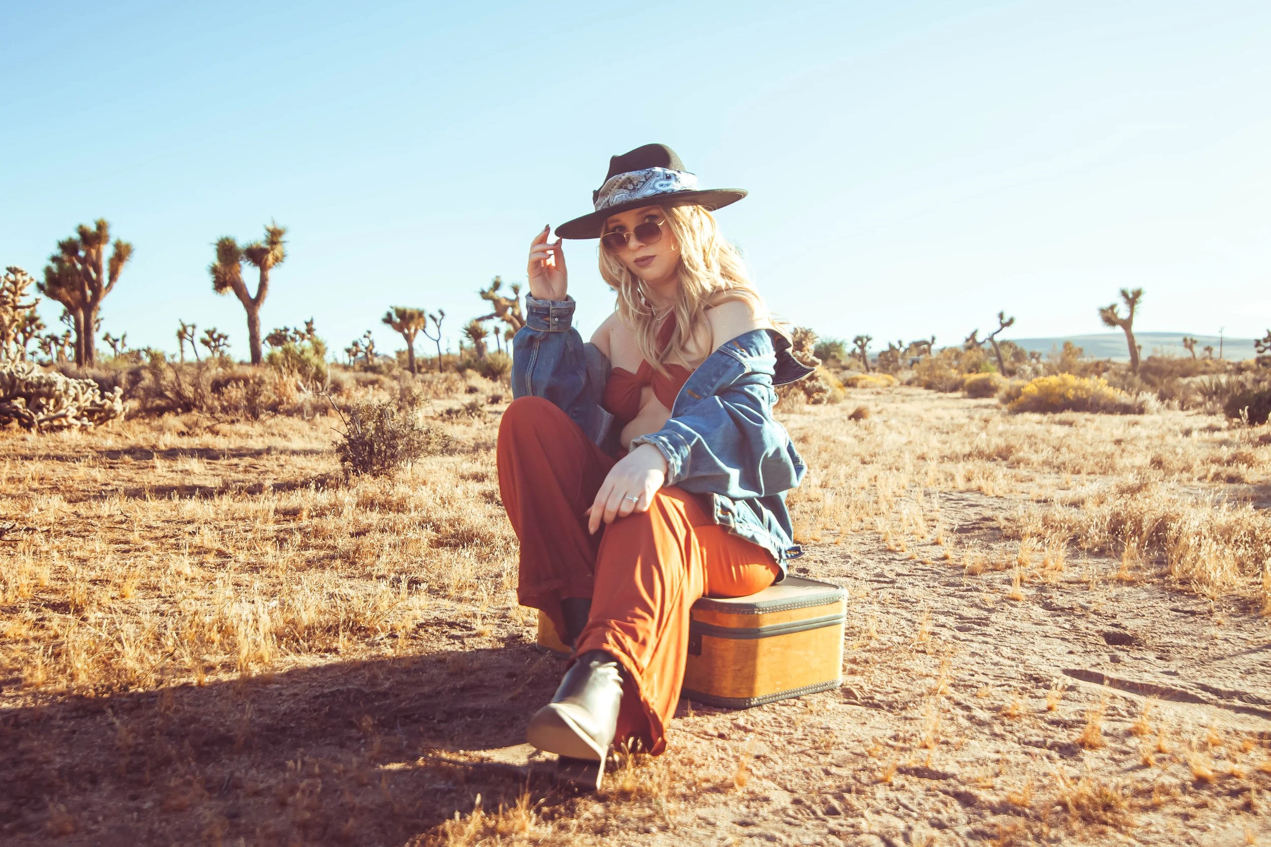 A woman sitting on a vintage suitcase in a desert landscape with Joshua trees, wearing a wide-brimmed hat, sunglasses, a denim jacket, and rust-colored pants.
