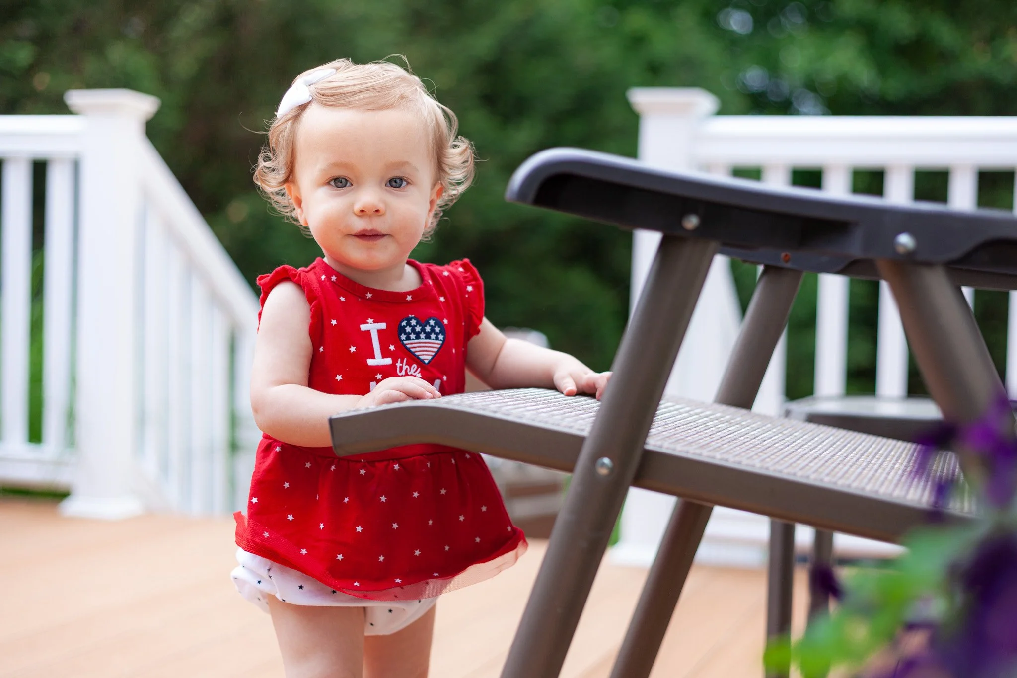 Candid children's portrait of a toddler girl with curly blonde hair and a white bow, wearing a red dress with an American flag heart, standing outside on a wooden deck with a white railing. She stares calmly and curiously into the camera.