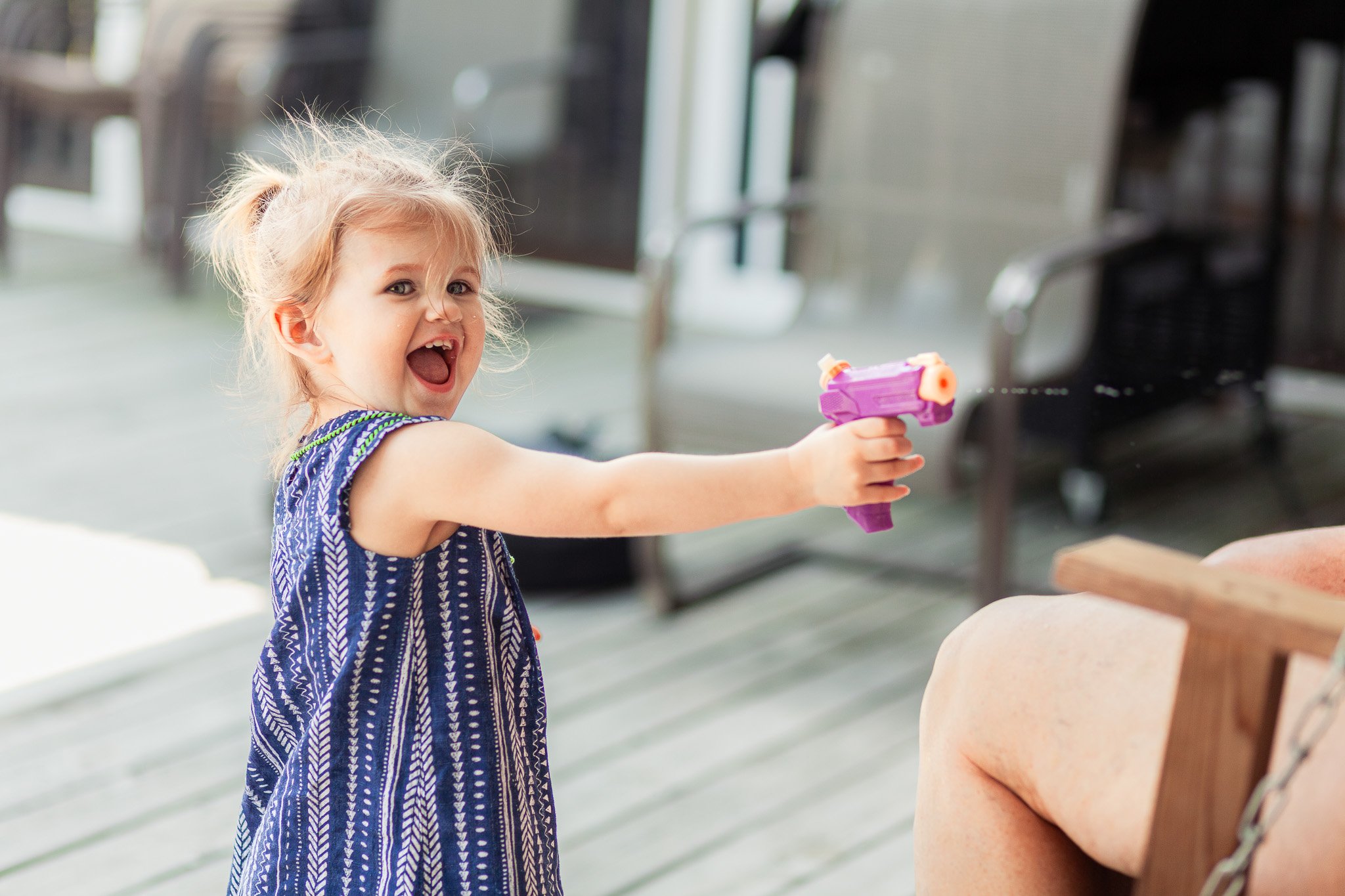 Young girl with blonde hair in a ponytail is smiling and playing with a purple water gun on a porch, aiming it at a person sitting on a swing.