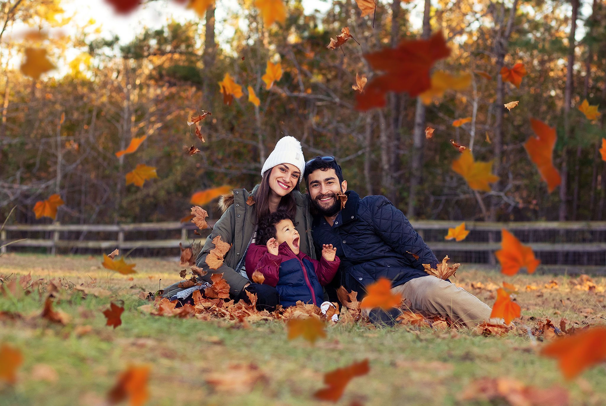 A  candid portrait of a family of three, two adults and one child, sitting on the ground smiling while autumn leaves swirl around them at a park during the fall season. 