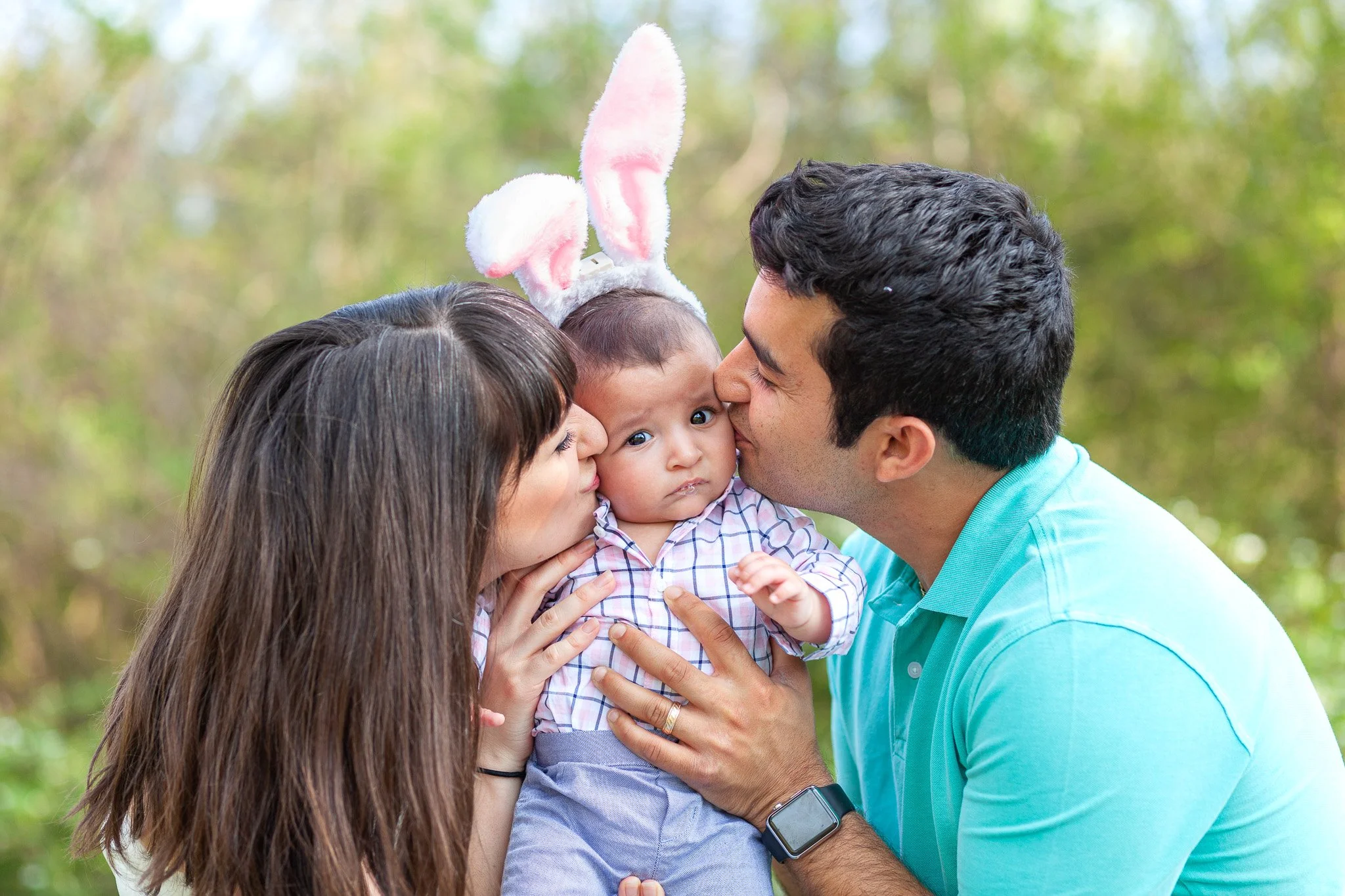 A family of three outdoors: a woman and a man kissing a young child wearing bunny ear headband.