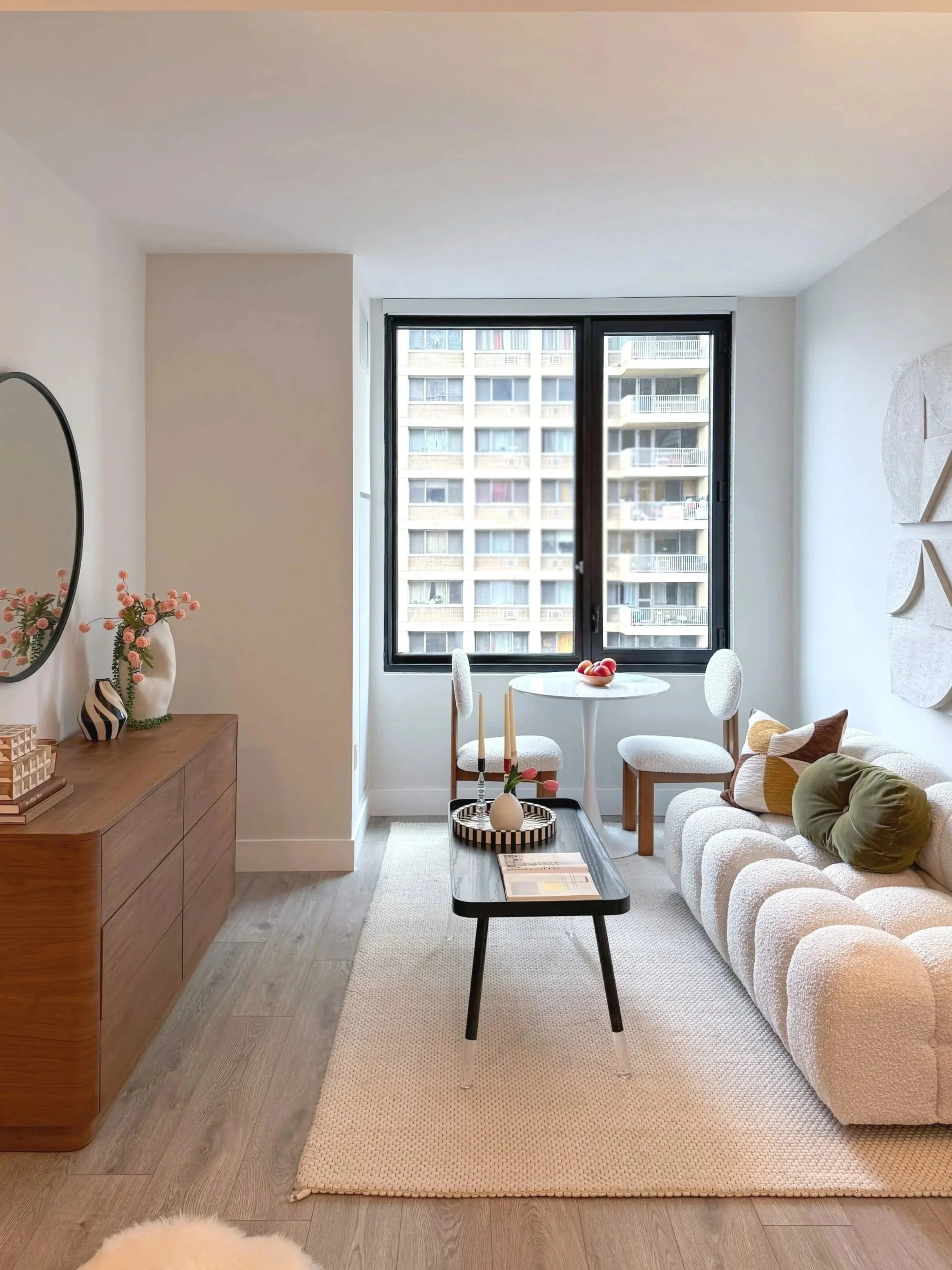 A modern living room with a large window showing a high-rise building outside. The room features a white textured sofa with colorful throw pillows, a black coffee table with decorative items, a round dining table with two white chairs, and a wooden sideboard with flower decorations.