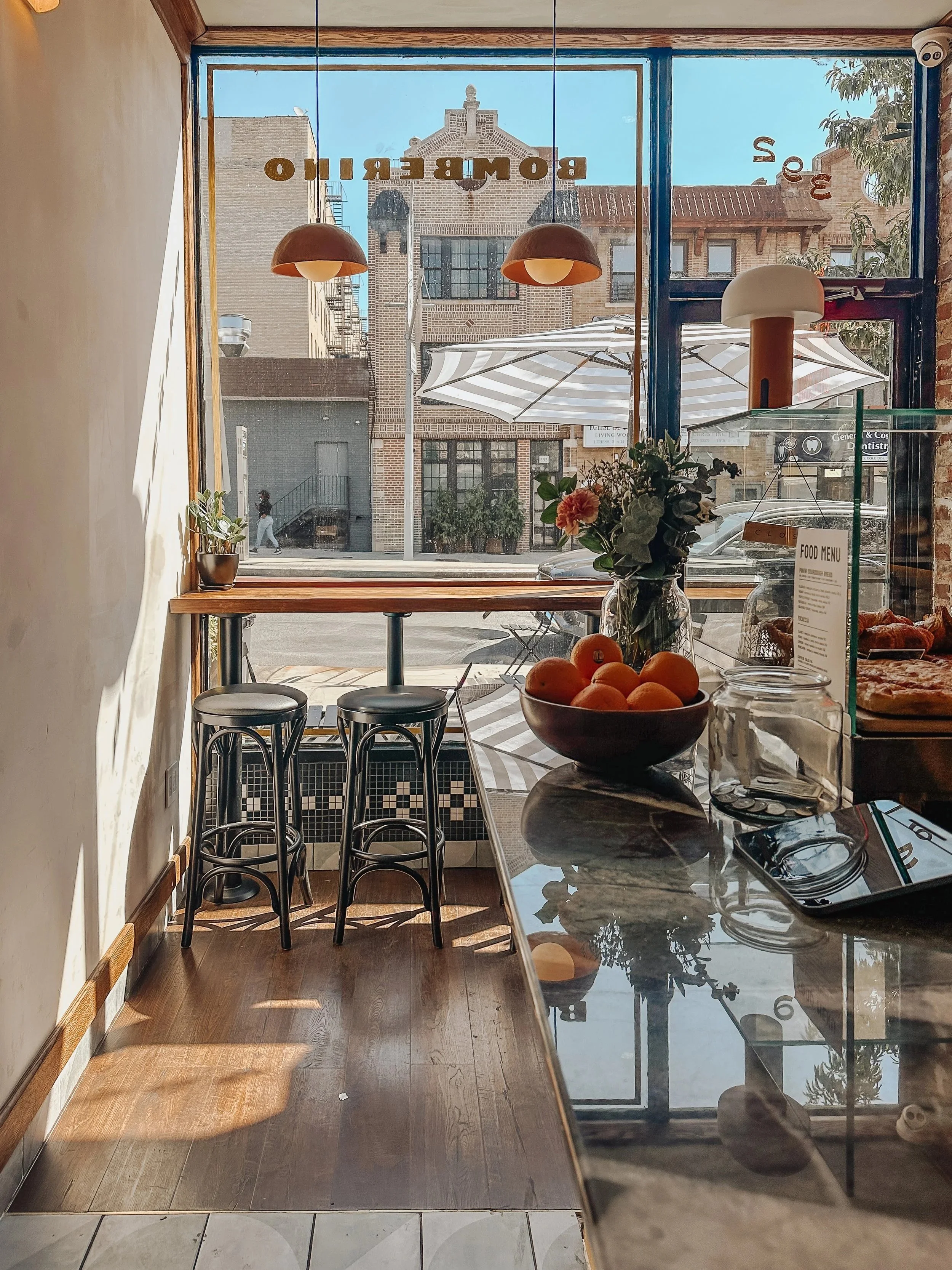 View from inside a cafe looking out the large front window, with three black bar stools at a high wooden counter. Outside, a building with a clock tower can be seen, along with a patio umbrella and parked cars. Inside, there is a bowl of oranges and a vase with flowers on the counter, with sunlight casting shadows on the wooden floor.