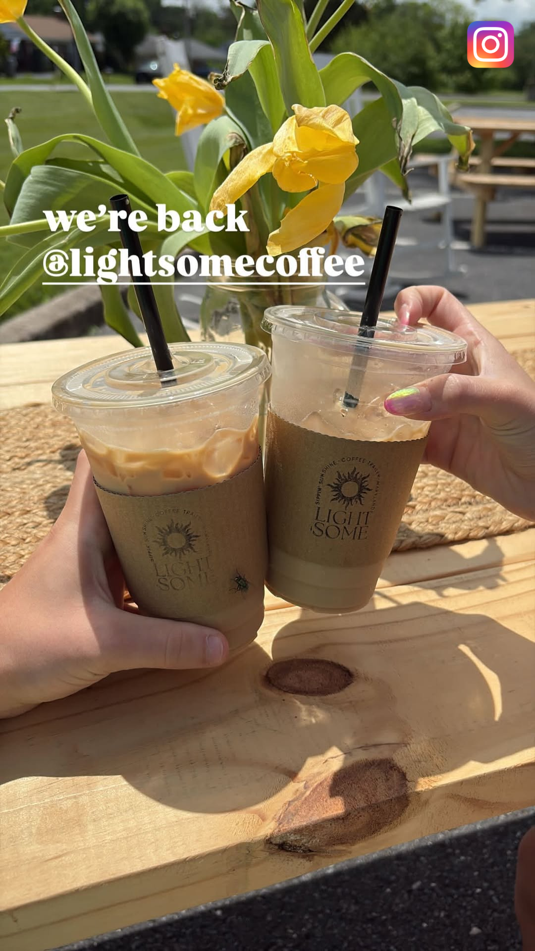 Two iced coffee drinks in to-go cups with light brown sleeves on a wooden table outdoors, with a background of yellow flowers in a vase and parked cars.