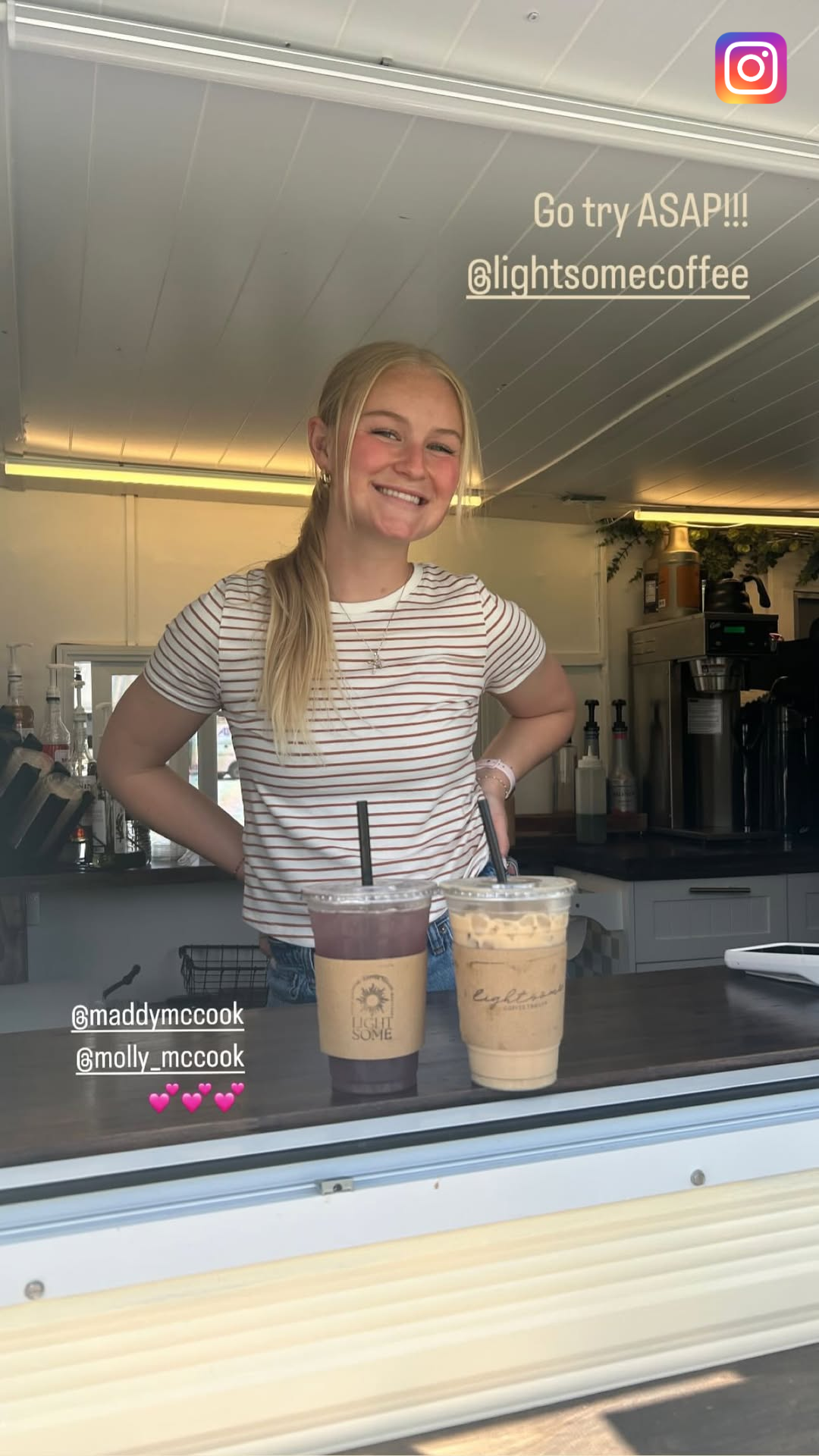 A young woman behind a coffee stand, smiling with hands on hips, has two iced drinks on the counter, and the background features a coffee station with equipment and supplies.