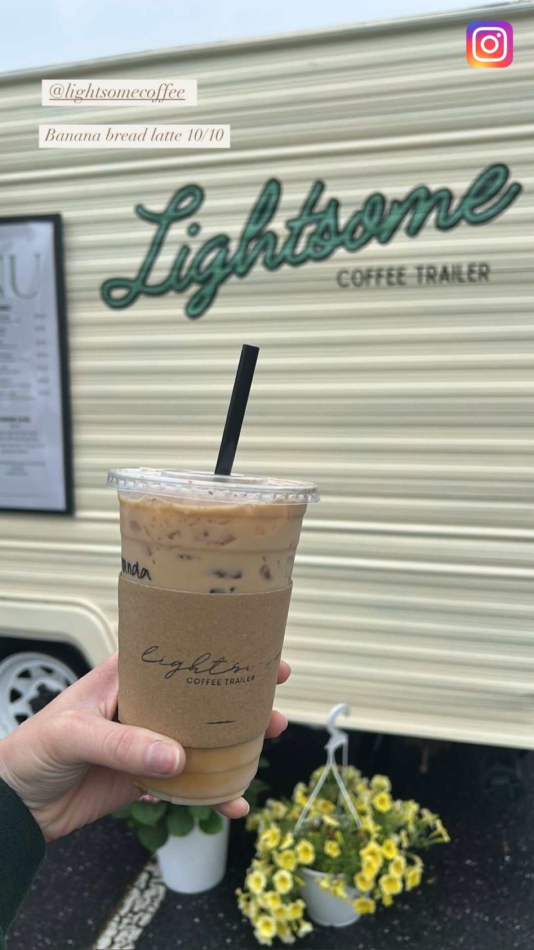 Hand holding a cold banana bread latte from Lightsome Coffee Trailer in a clear plastic cup with a black straw, in front of a beige mobile coffee trailer with the Lightsome logo and a flower pot with yellow flowers.