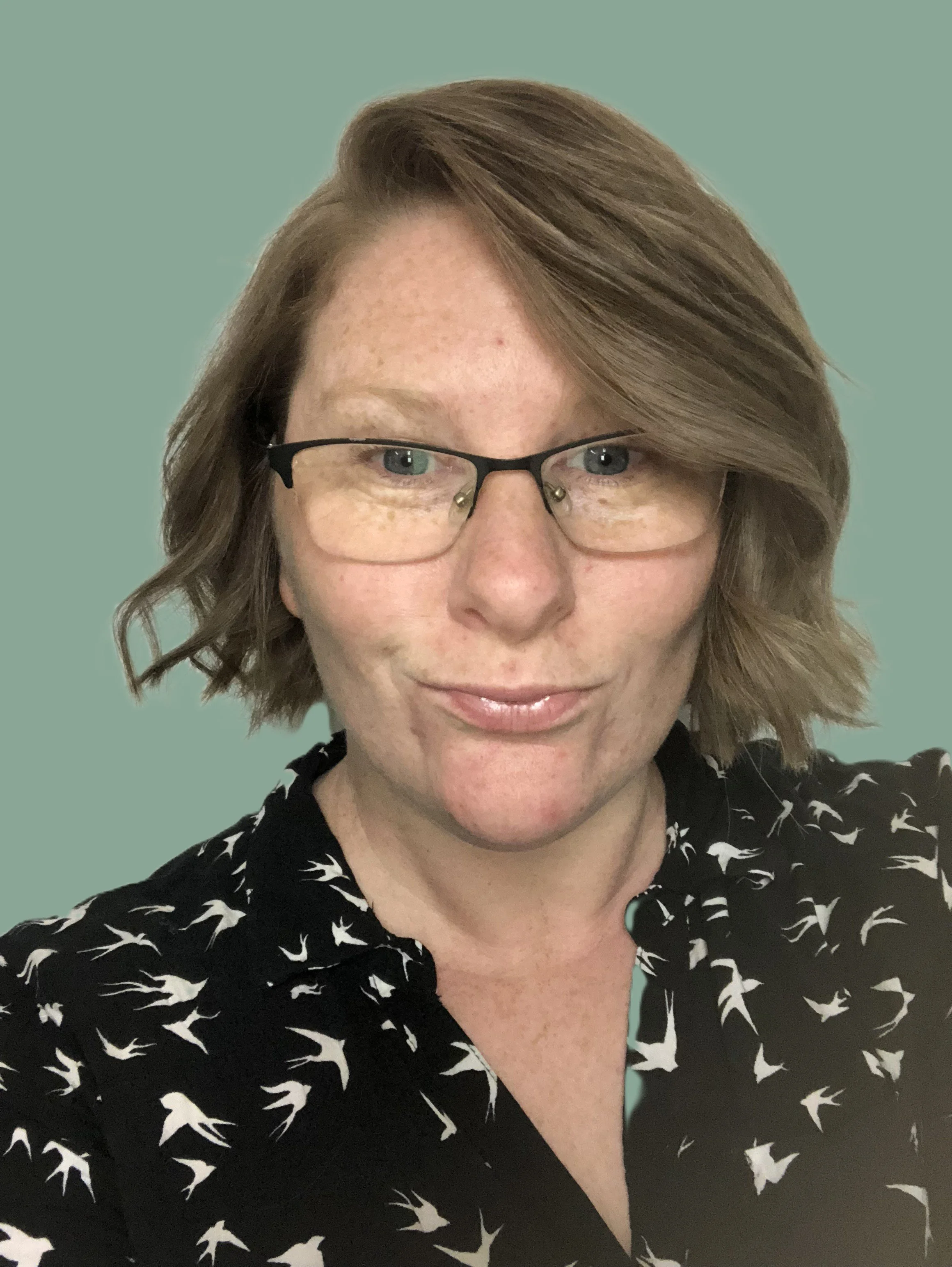 In image: founder of pragmatic spark collective, Kathryn, is wearing glasses and has short wavy hair. Kathryn is wearing a black collared shirt with small white bird details. The background is green.