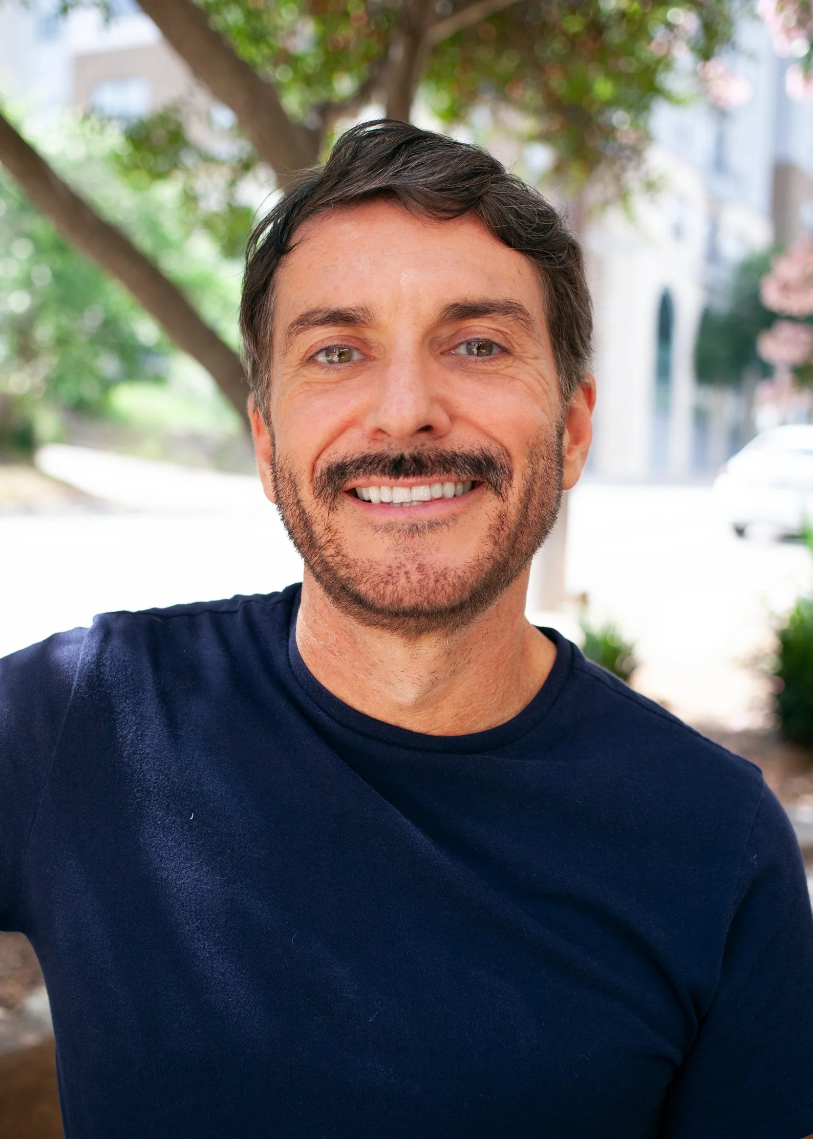 A smiling male massage therapist with dark hair, a beard, and light-colored eyes, wearing a navy blue shirt, standing outdoors with green trees and blurred city buildings in the background.