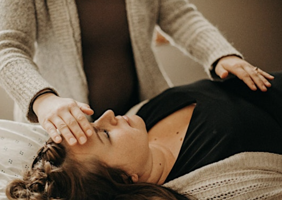 A woman lying on her back with eyes closed, receiving a gentle head massage from another person, in a relaxed setting.