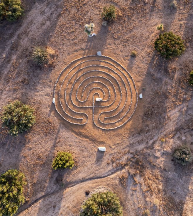 An aerial view of a chalk maze on dirt ground, surrounded by trees and small structures.
