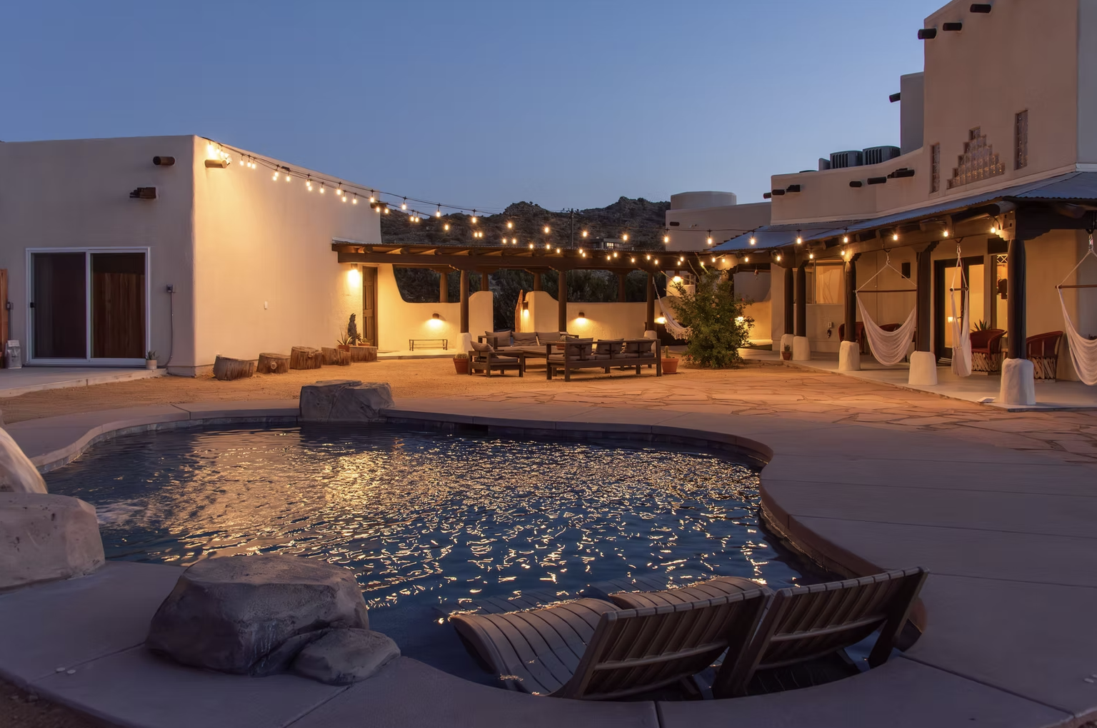 An outdoor patio area at dusk with a small swimming pool reflecting string lights overhead, surrounded by wooden furniture, hammocks, and desert-style houses with mountain views in the background.