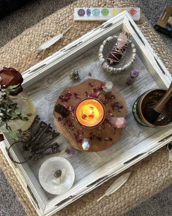 A white distressed tray contains a lit candle, crystals, a bundle of sage and palo santo, an essential oil roller, a stack of prayer or meditation beads, a bundle of keys, and a small bowl with a candle inside on a woven surface.